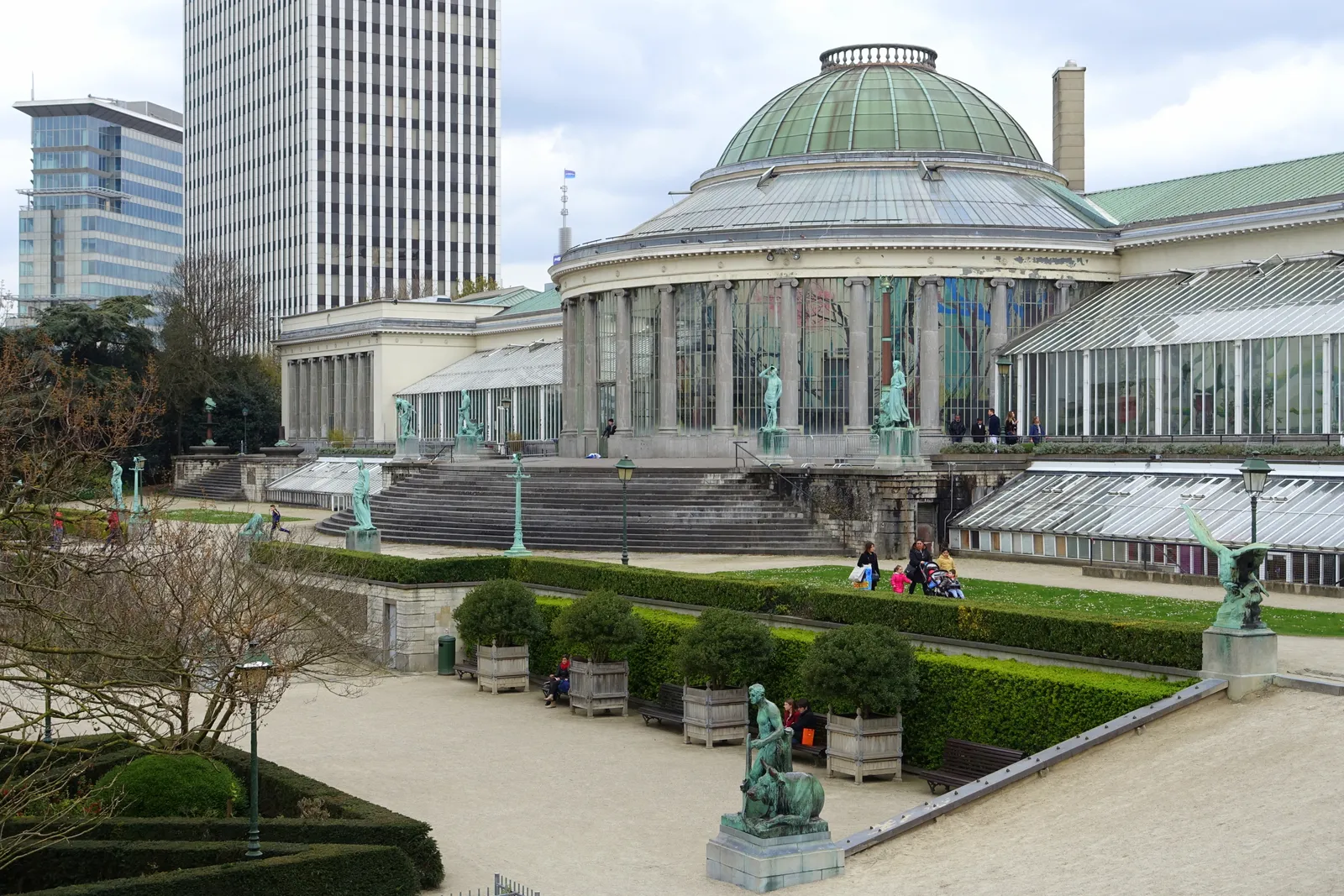 The domed glasshouse rotunda of the Botanique in Brussels above the terraced botanical garden, with bronze statues along the clipped hedges and office towers behind