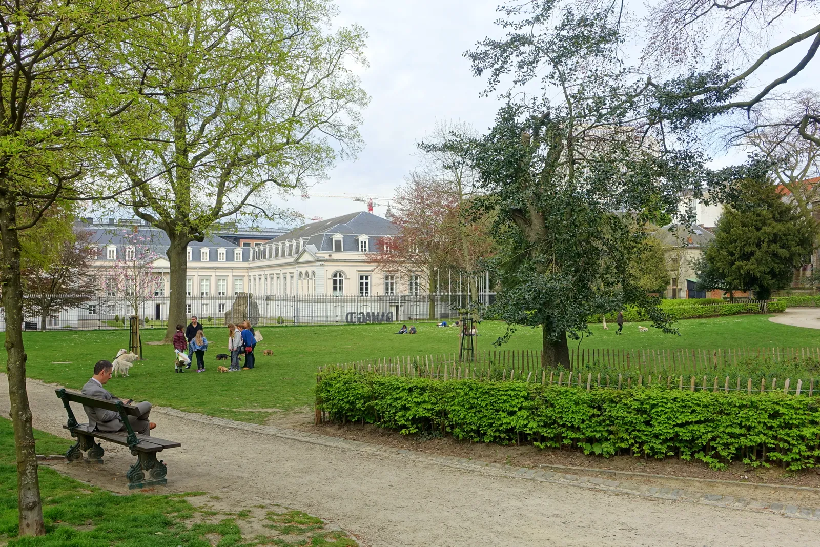 Lawns and bare spring trees in the Egmont Park in Brussels, with people sitting on benches along a gravel path and the long white front of the Egmont Palace behind