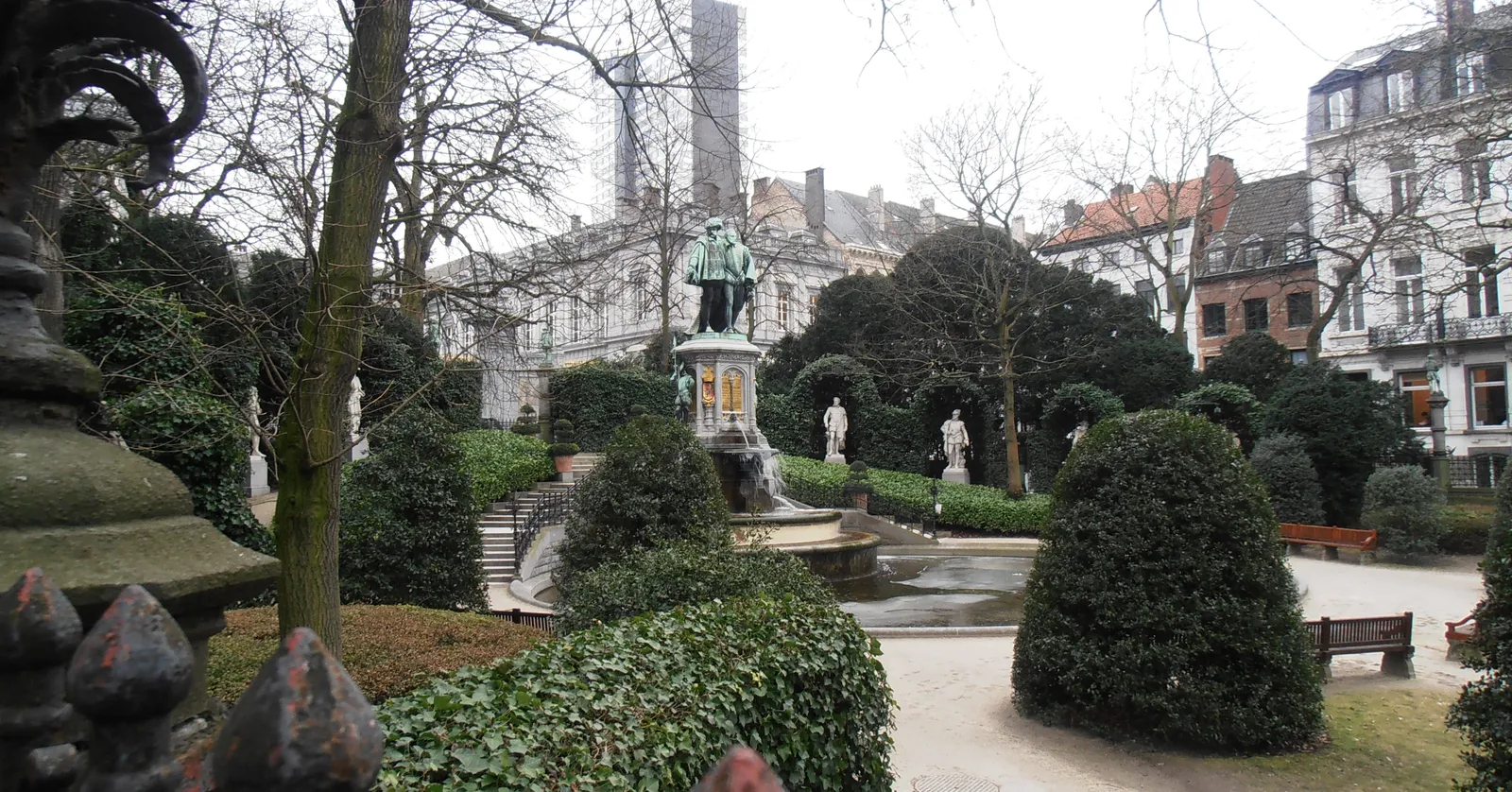 The garden at the Square du Petit Sablon in Brussels seen through its ornate railing, clipped hedges around a fountain and monument with small bronze figures on columns along the far side