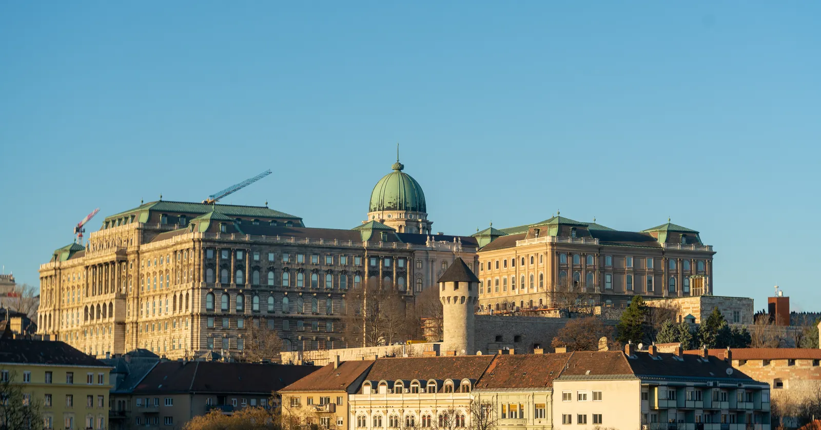 Buda Castle in Budapest seen from across the Danube in low sun, a long ochre palace range with green copper roofs and a green dome over the centre, standing on the hill above the rooftops of the Víziváros district