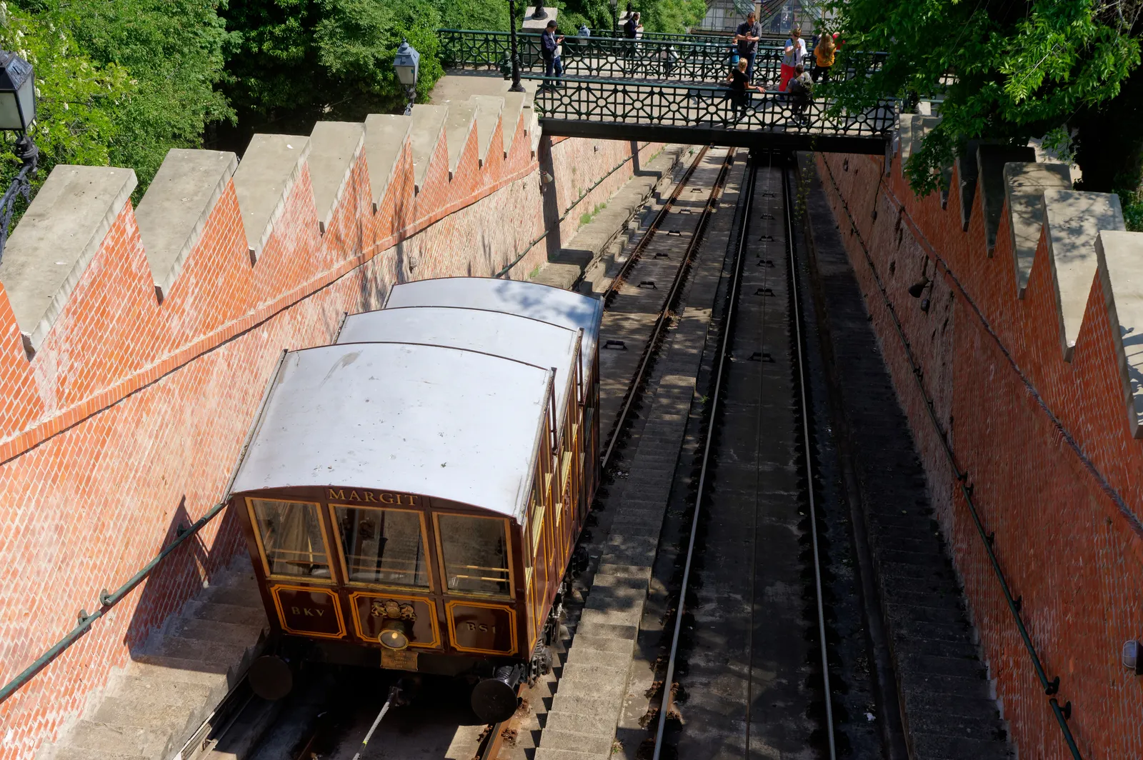 A wooden funicular carriage climbing the track cut into Castle Hill in Budapest between brick retaining walls, with an iron footbridge crossing above it and people looking down from the parapet