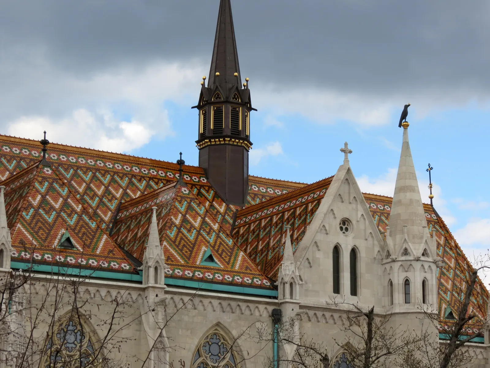 The roof of Matthias Church in Budapest, covered in diamond-patterned glazed tiles in orange, green and white, with a slender dark spire and carved white stone gables rising above it