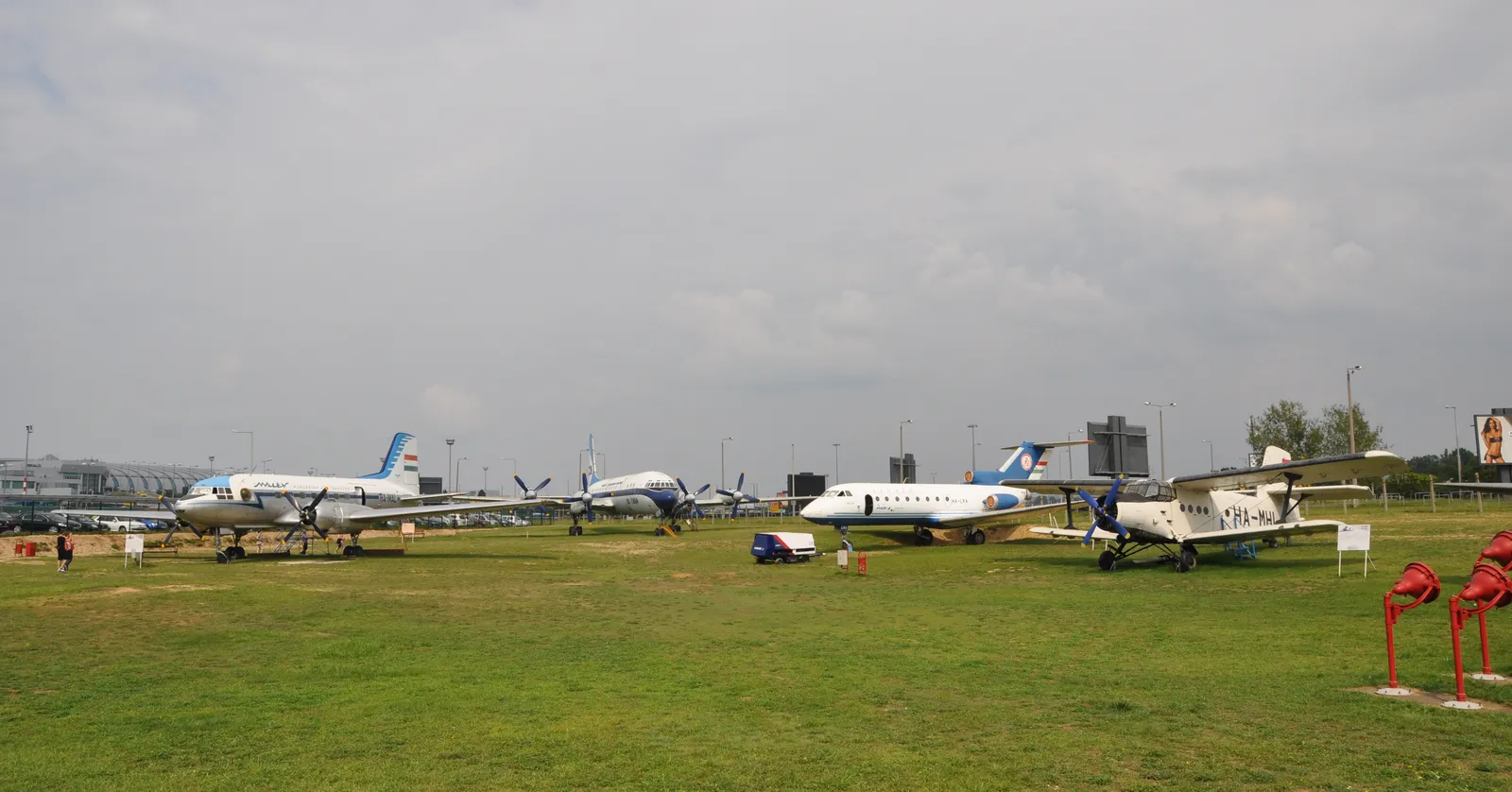 A line of retired airliners parked on grass at the Aeropark museum beside Budapest airport: a Malév propeller aircraft, a four-engined turboprop, a small white jet and a blue and white biplane, with the terminal roofline in the distance