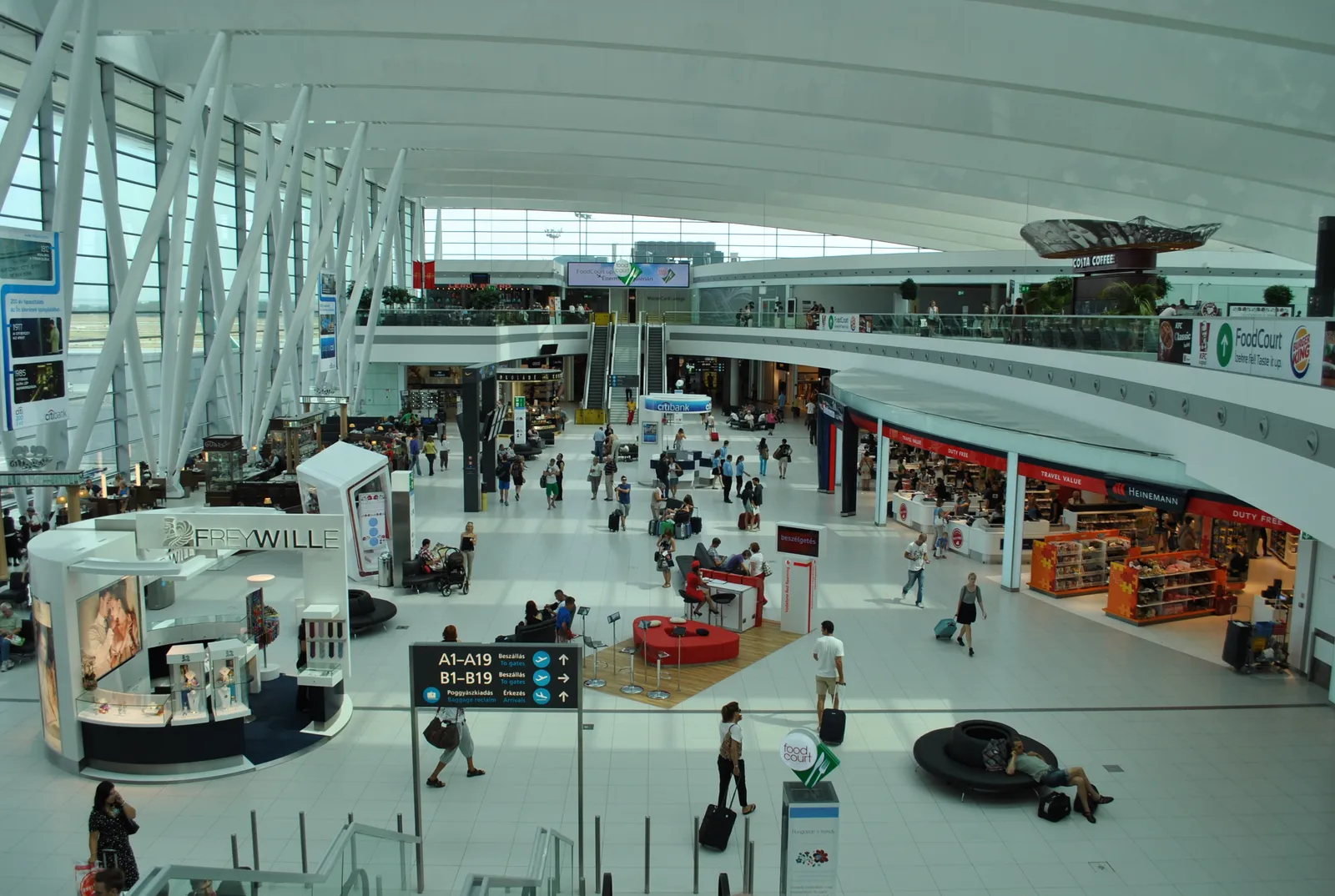 The SkyCourt hall at Budapest airport seen from an upper level, a wide white space with diagonal steel struts along a glazed wall, shops and cafés around the edges and passengers crossing the floor below