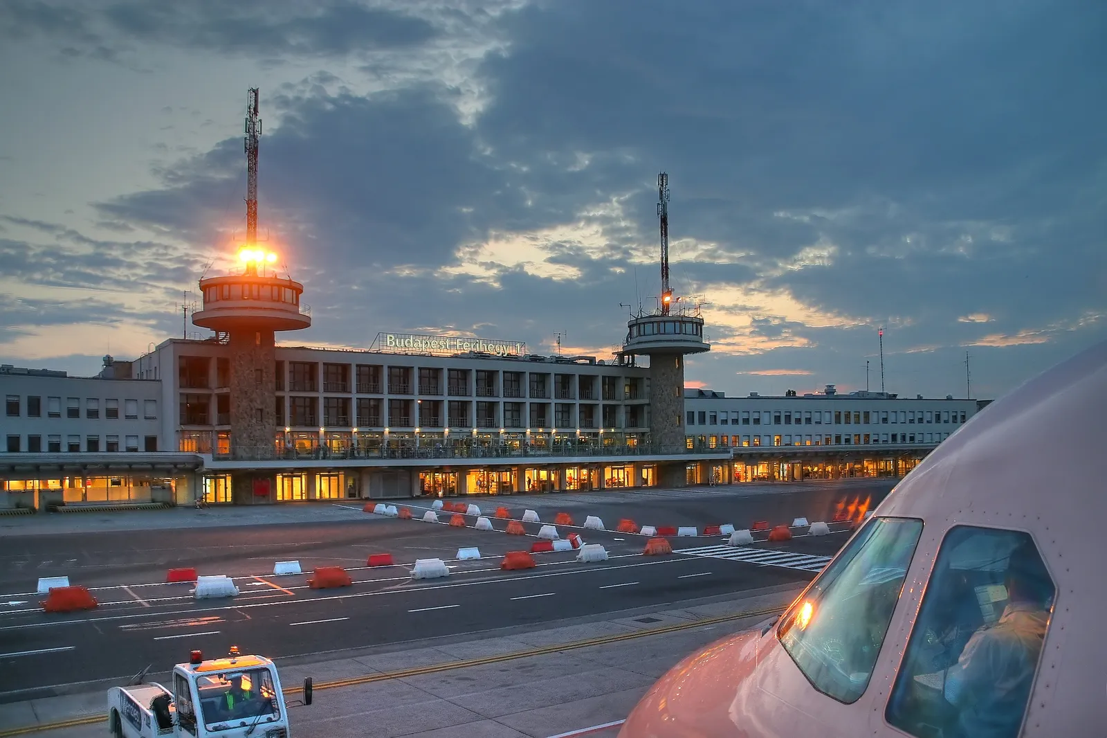 The disused Terminal 1 building at Budapest airport at dusk, a long low modernist block lit from within, with a stone-clad control tower at each end and an aircraft nose in the foreground
