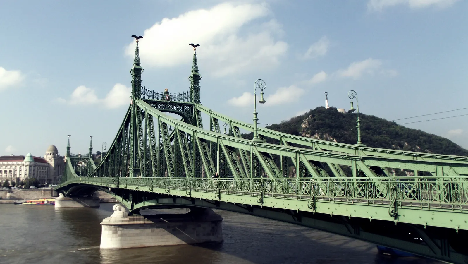 Liberty Bridge in Budapest seen from the Pest bank, a pale green painted iron truss bridge with decorated masts rising from its piers, crossing the Danube towards the wooded slope of Gellért Hill