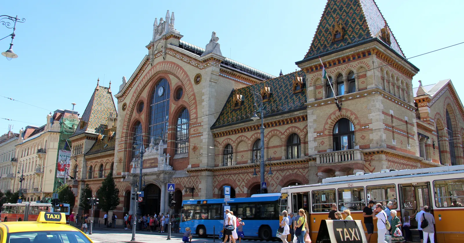 The Great Market Hall in Budapest from Fővám tér, a wide brick and stone gable with a rose window and a steep roof of patterned glazed tiles, with trams, a bus and taxis passing in front of it