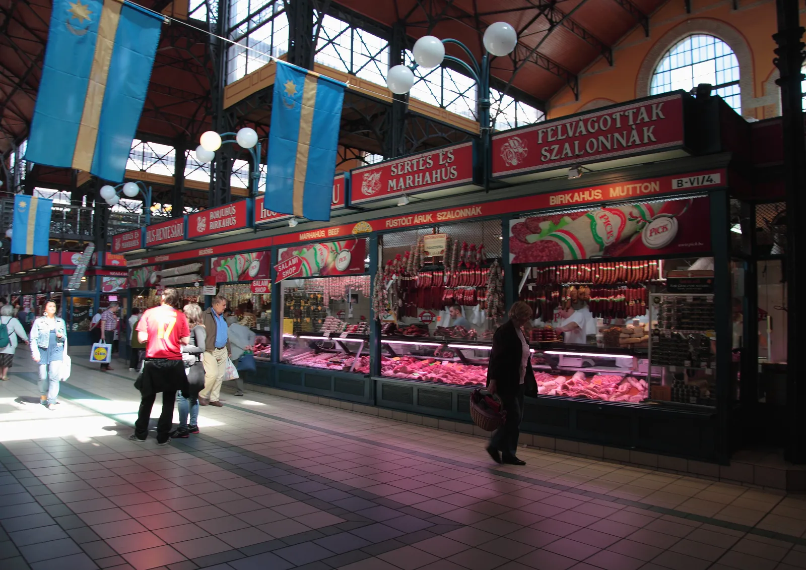 Inside the Great Market Hall in Budapest, a row of red-fronted butchers' counters hung with sausages and salami under the iron roof structure, with shoppers walking the wide tiled aisle in front