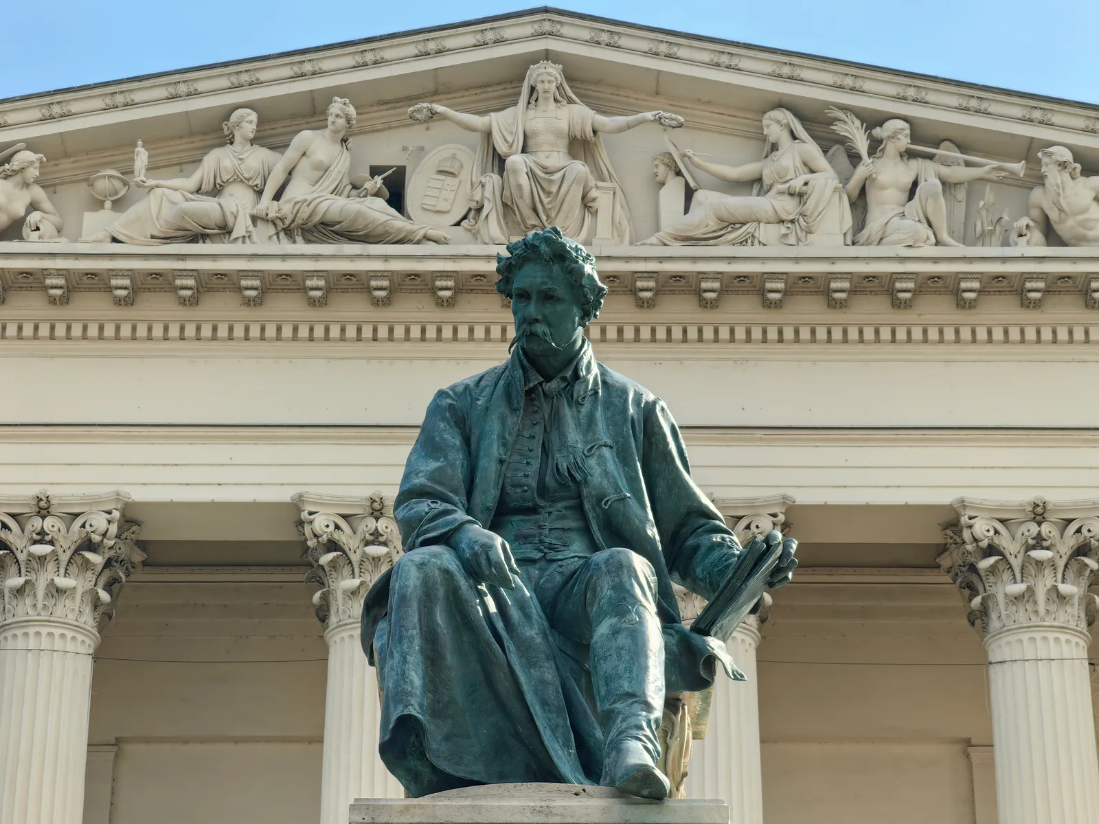 The portico of the Hungarian National Museum in Budapest, Corinthian columns below a carved stone pediment full of figures, with a seated bronze statue of the poet János Arany on a plinth in front