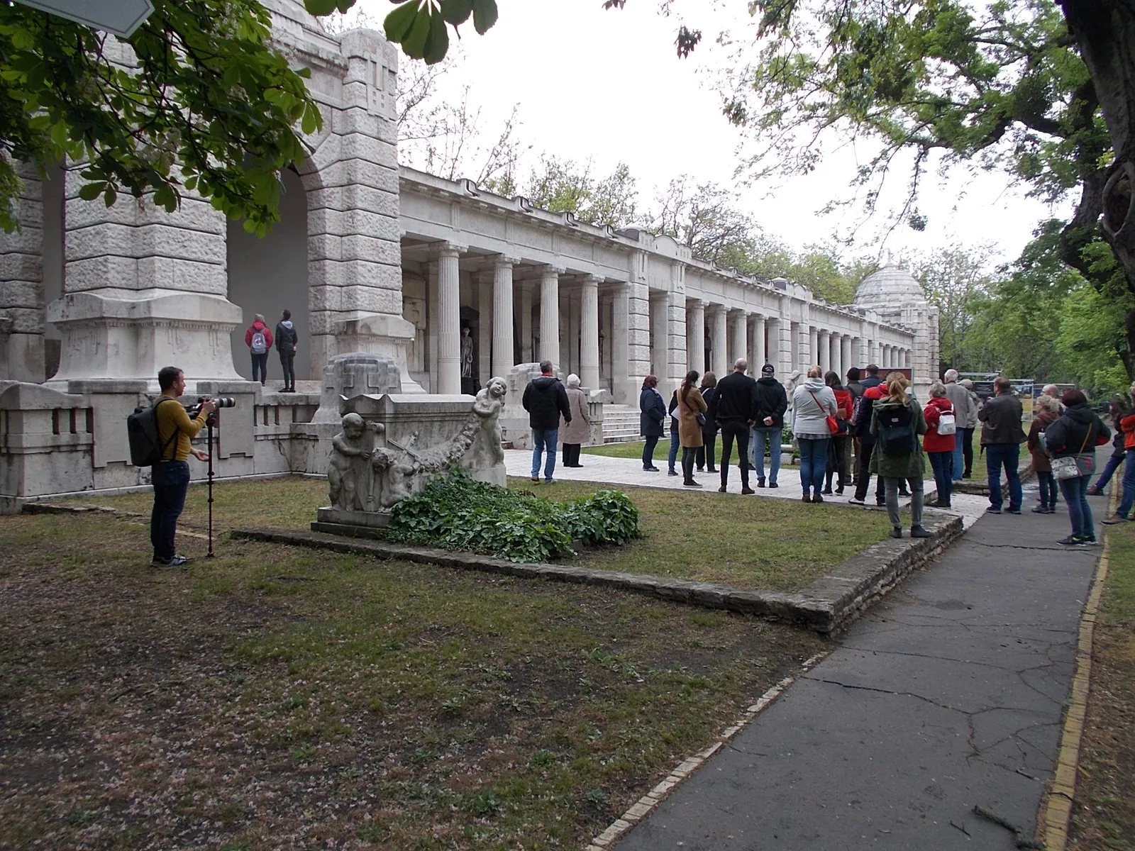 The stone arcades at the Fiumei road graveyard in Budapest, a long colonnade of family tombs under a covered walkway, with a small group of visitors standing in front of it