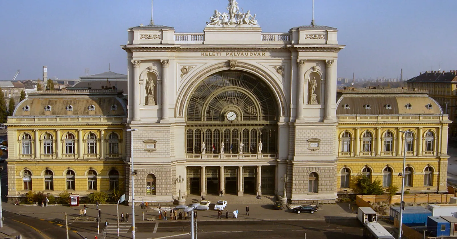The front of Budapest Keleti railway station seen from across the square, a pale stone triumphal arch filled with an iron and glass fanlight, KELETI PÁLYAUDVAR carved above it and a sculptural group on the roofline, with yellow wings on either side