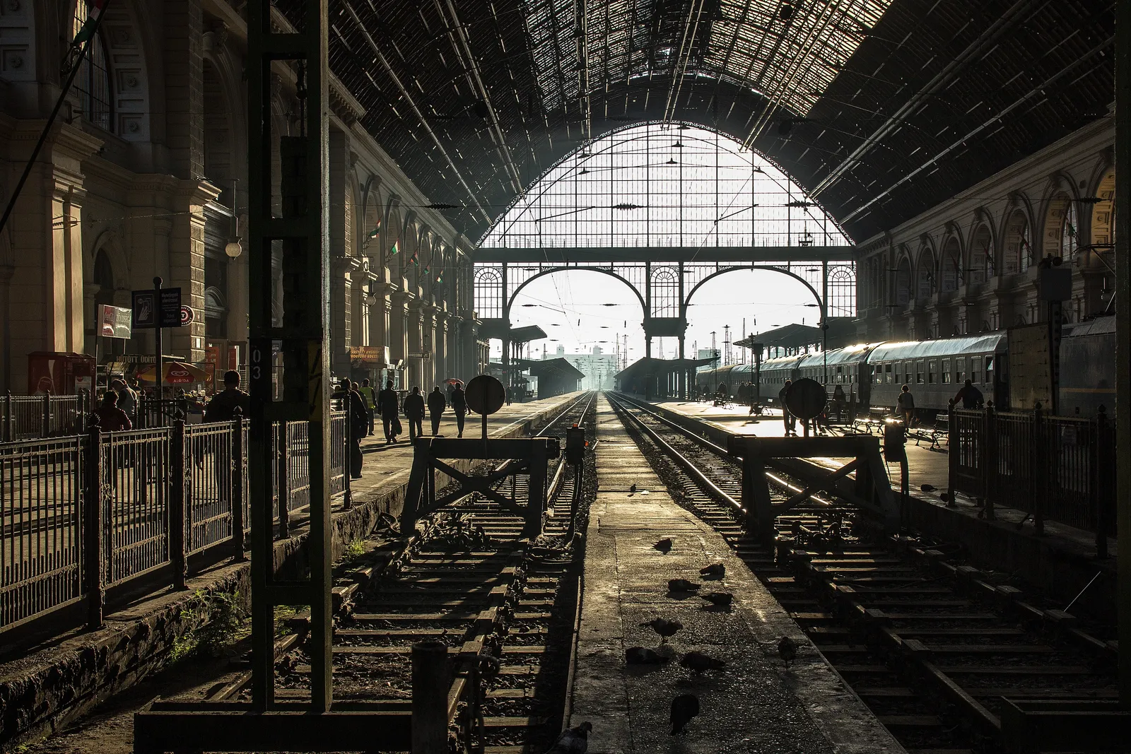 Inside the train shed at Budapest Keleti station, looking down the tracks between platforms towards the tall arched glass end wall with daylight coming through it