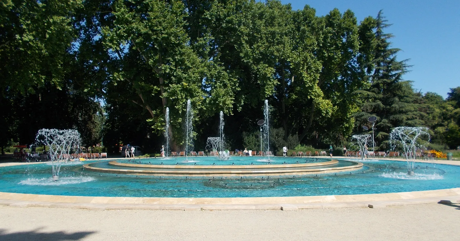 The musical fountain at the southern end of Margaret Island in Budapest, a wide circular pool of turquoise water with jets rising from concentric stone rings, benches and tall plane trees behind it
