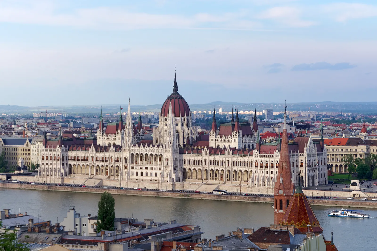 The Hungarian Parliament Building in Budapest seen from across the Danube, a long white neo-Gothic frontage of spires and arcades along the water with a red dome rising over the centre