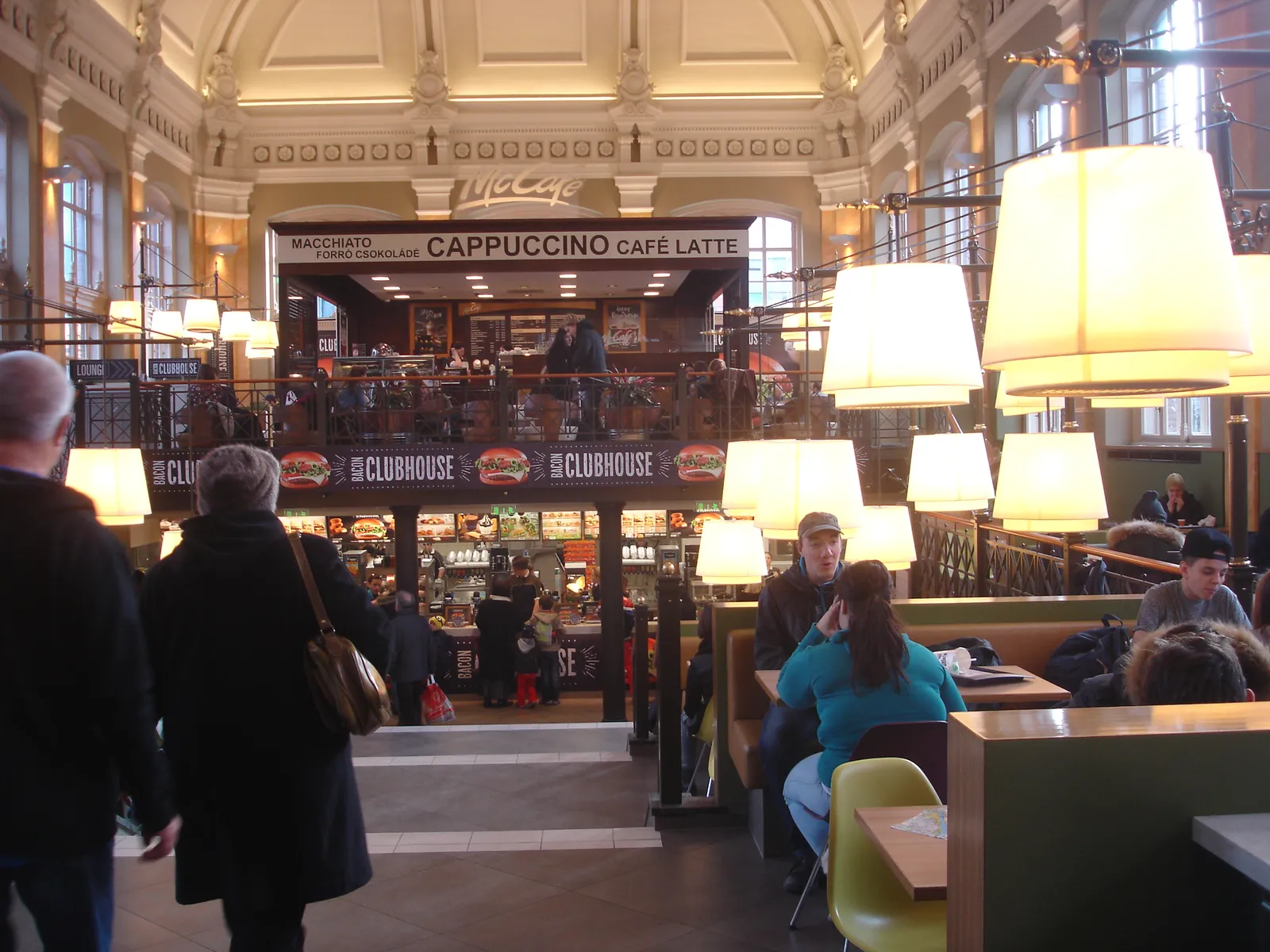 The former station dining room at Budapest Nyugati, a high hall with a moulded plaster ceiling, a galleried upper floor behind iron railings and hanging lamps over the tables, now in use as a fast-food restaurant