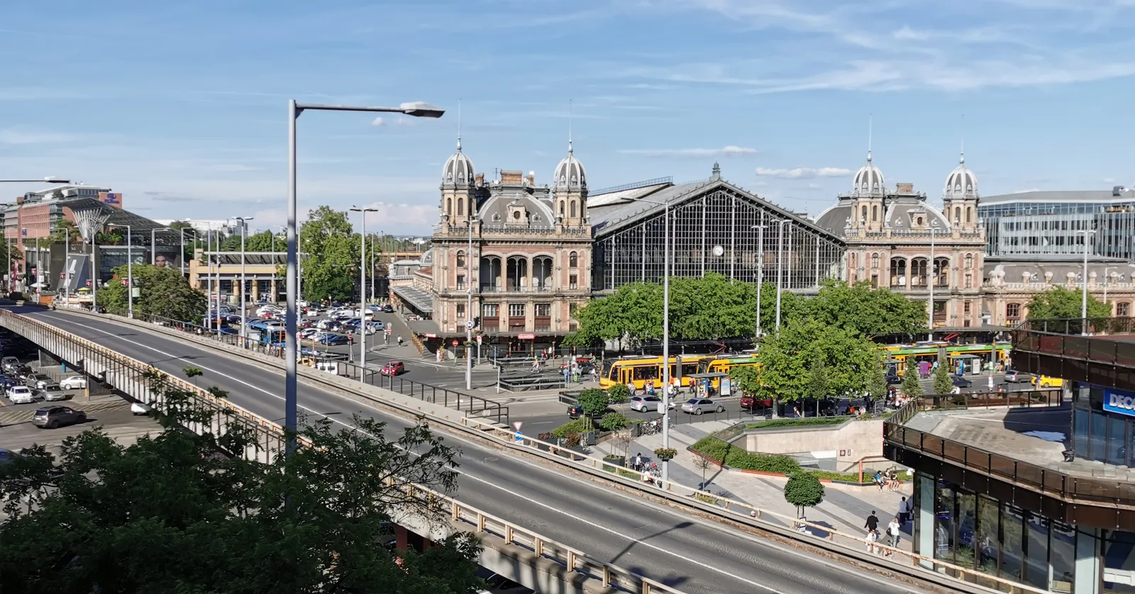Budapest Nyugati railway station seen from a raised road, its wide iron and glass gable end flanked by two domed brick pavilions, with yellow trams and trees along the boulevard in front of it
