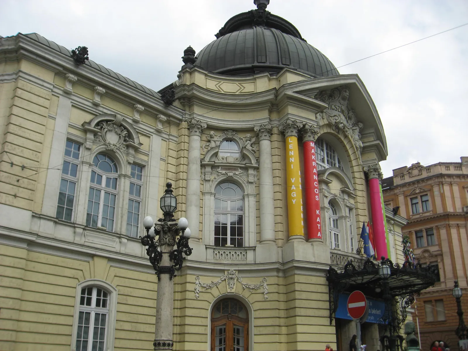 The front of the Comedy Theatre in Budapest, a pale yellow neo-baroque building with columns, carved reliefs and a dark dome above it, hung with printed banners between the upper windows