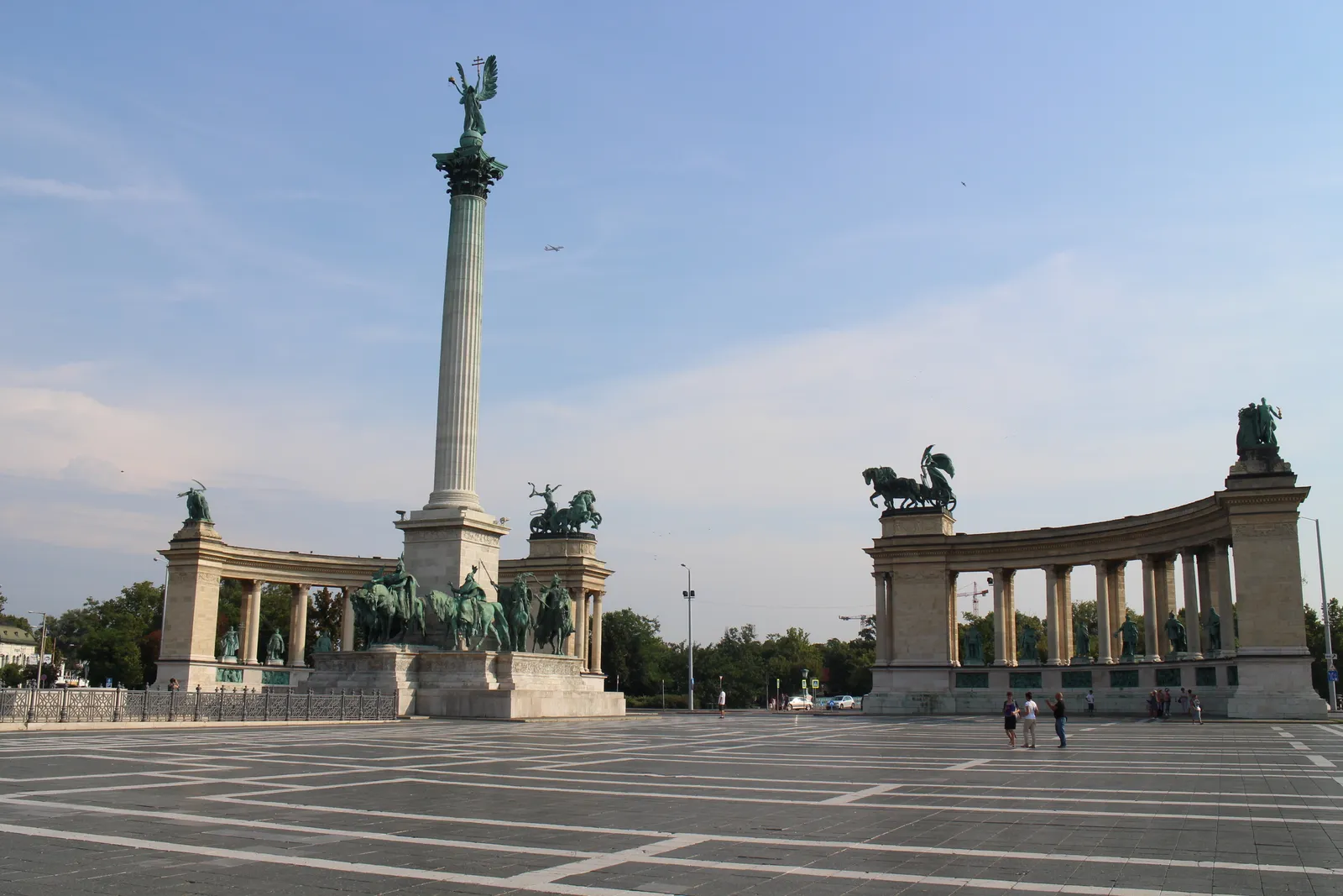 Heroes' Square in Budapest, a wide empty paved square with a tall stone column carrying a winged figure at its centre, bronze horsemen at the base and two curved colonnades of statues on either side