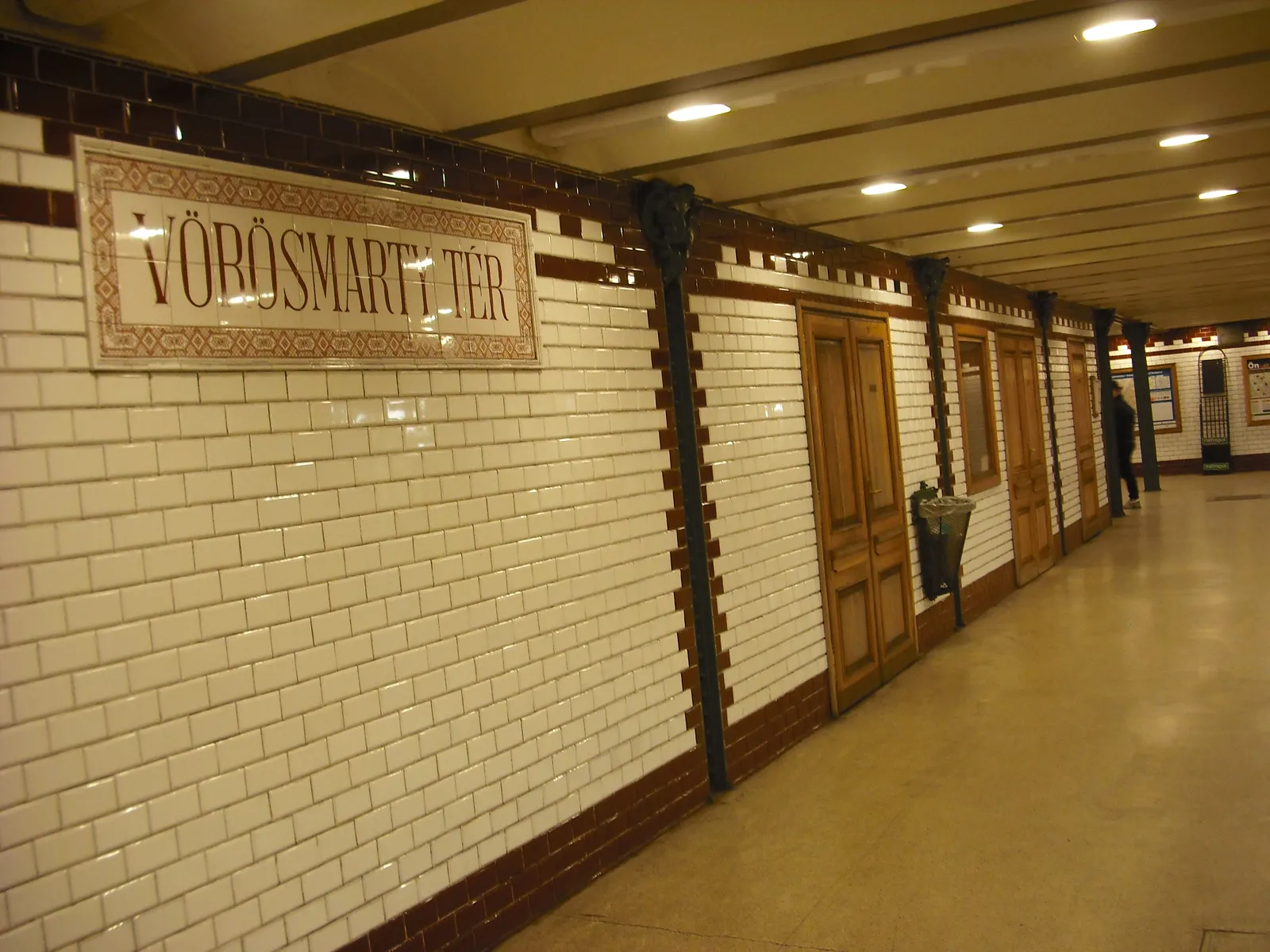 A platform on Budapest's Millennium Underground, lined in white tiles with a brown tile band, cast-iron columns holding a low coffered ceiling, wooden doors along the wall and a tiled station nameplate