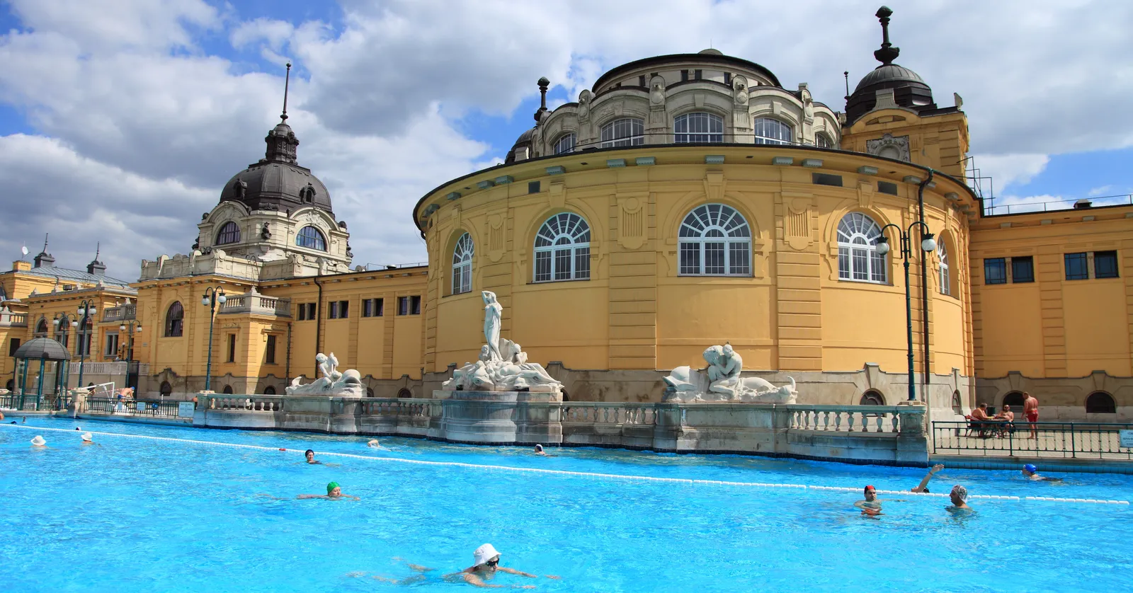 The main outdoor pool at Széchenyi Baths in Budapest, bright blue water with swimmers in coloured caps, below a curved ochre neo-baroque bath building with domed roofs and white stone figures along the poolside balustrade