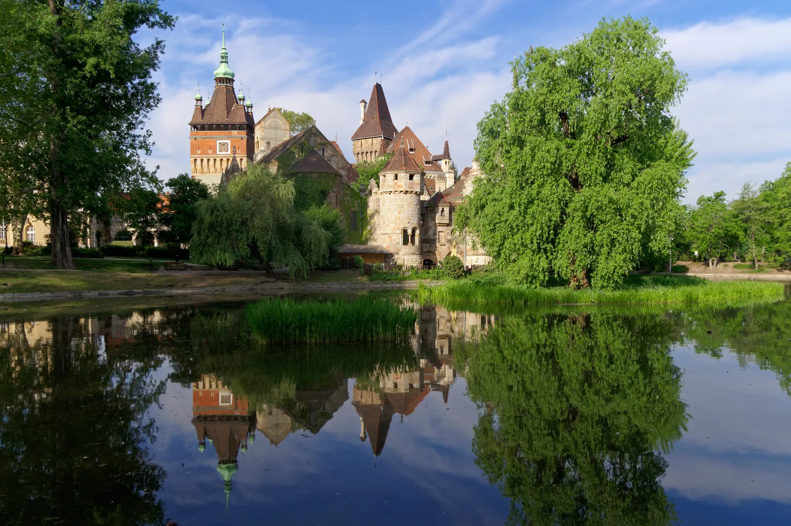 Vajdahunyad Castle in City Park, Budapest, its towers and steep tiled roofs rising behind willows and reflected in the flat water of the boating lake in front of it