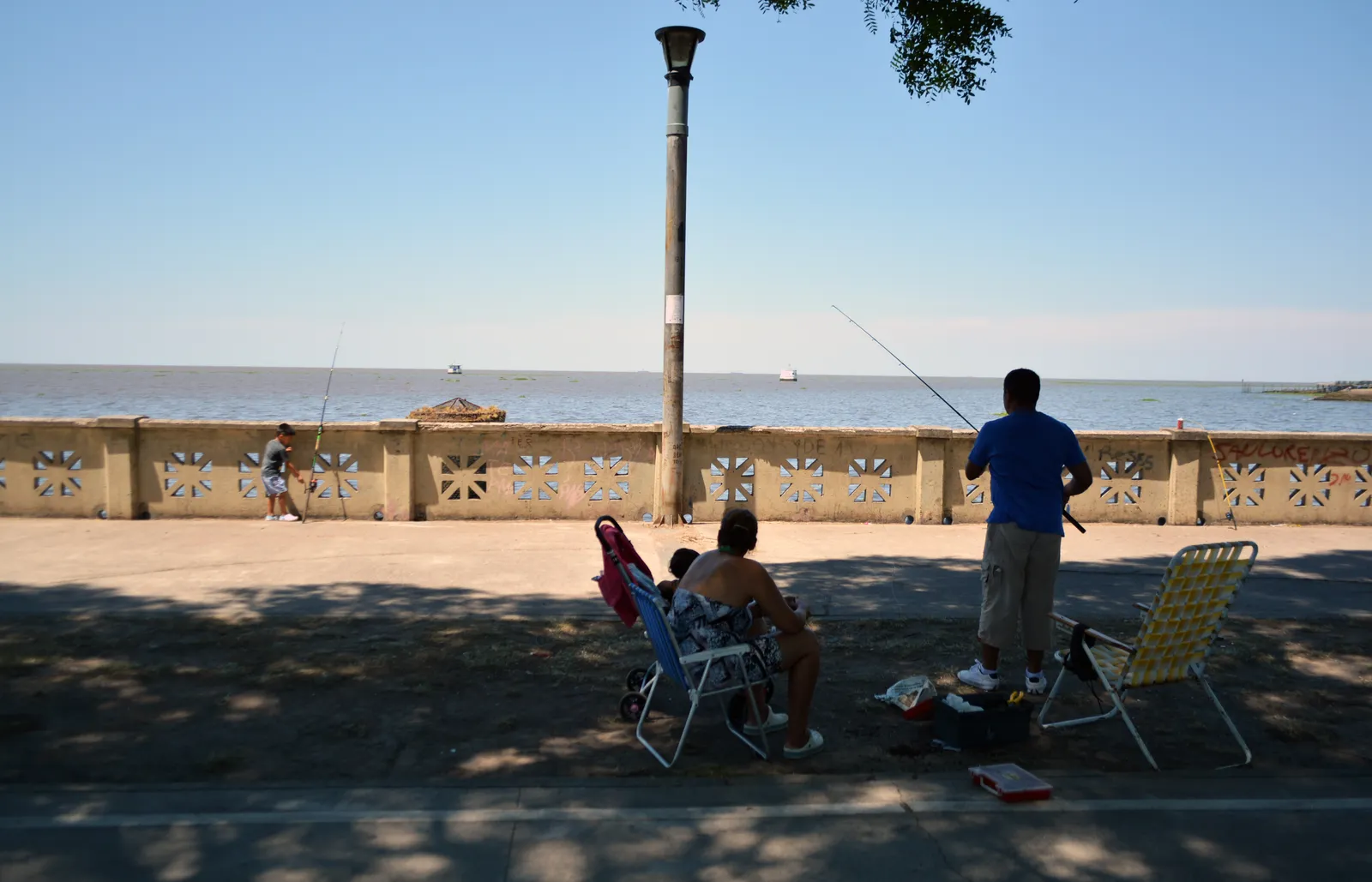 People fishing over the decorated stone balustrade of the Costanera Norte promenade in Buenos Aires, folding chairs on the path behind them and the brown Río de la Plata stretching to the horizon