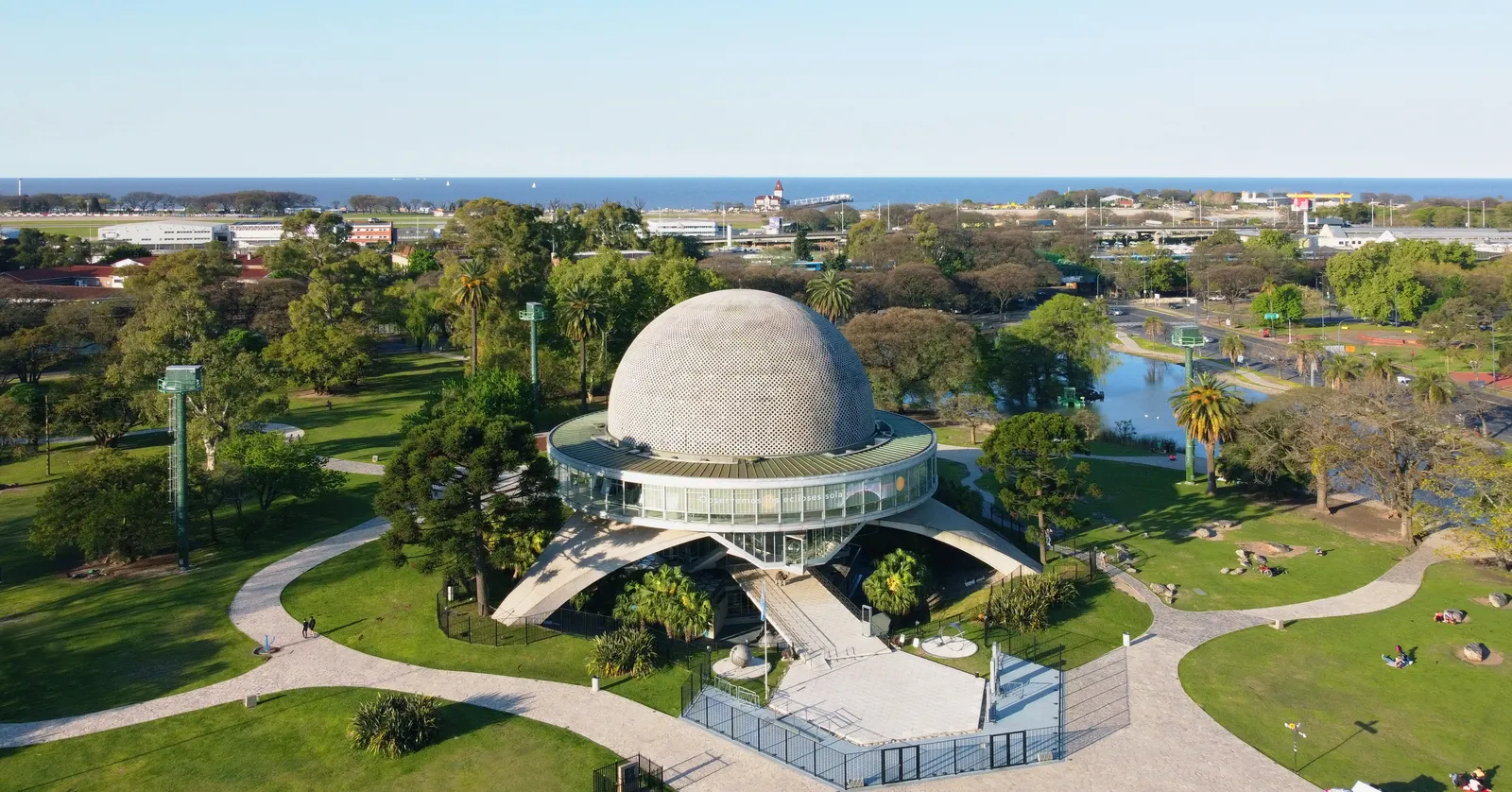 An aerial view of the Galileo Galilei planetarium in Buenos Aires, its perforated grey dome resting on three curved concrete arches above a glazed drum, with park lawns, gravel paths and a lake around it and the river beyond