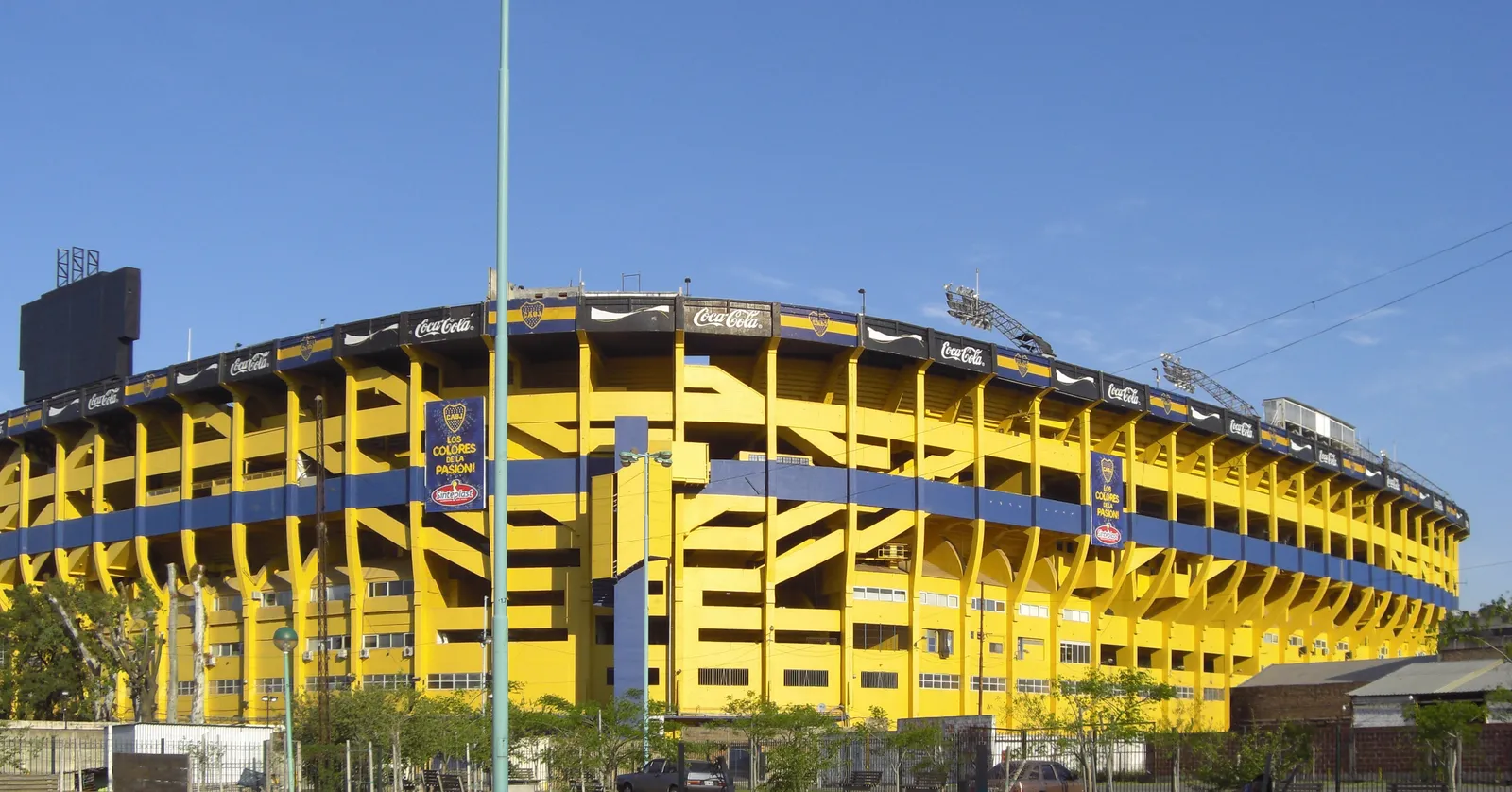 The outside of La Bombonera stadium in Buenos Aires, its steep concrete stands painted in bright yellow and blue stripes and curving away under a clear sky, with grass and a footpath in front