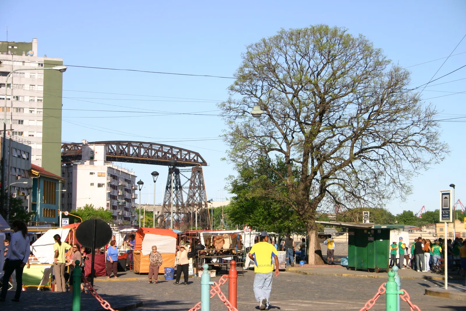 The steel lattice span of the Nicolás Avellaneda transporter bridge rising over the Riachuelo at the Vuelta de Rocha in La Boca, Buenos Aires, with market stalls and a large bare tree in the foreground