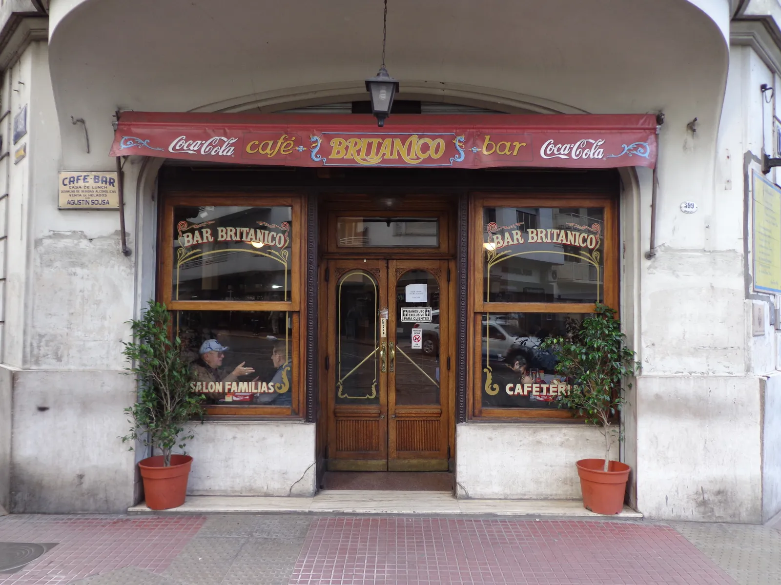 The street-level entrance of Bar Británico in Buenos Aires, its wooden doors and windows framed by a striped Coca-Cola awning reading Café Británico, with potted plants flanking the doorway