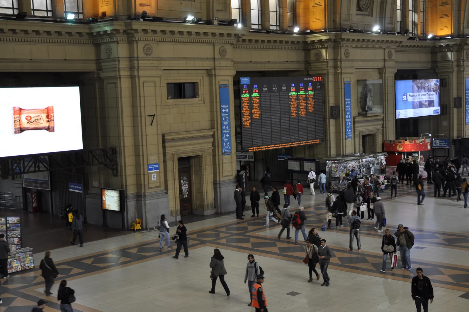 The wide interior concourse of Constitución station in Buenos Aires, with a departures board mounted high on a stone wall and passengers crossing the patterned floor below