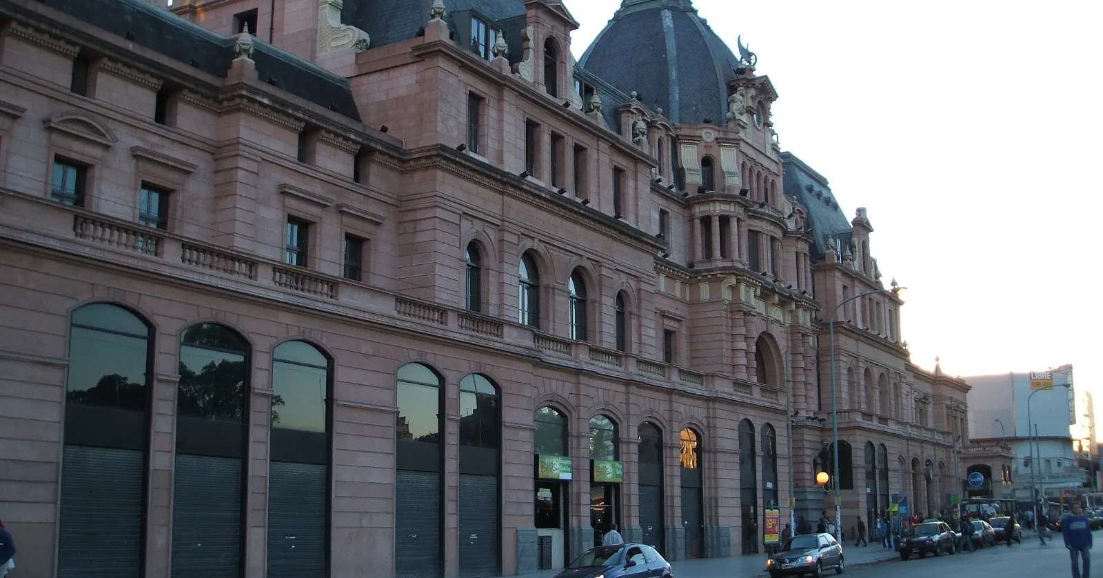The long pink stone facade of Constitución railway station in Buenos Aires at dusk, with a domed corner tower rising above rows of arched windows and cars parked along the street in front