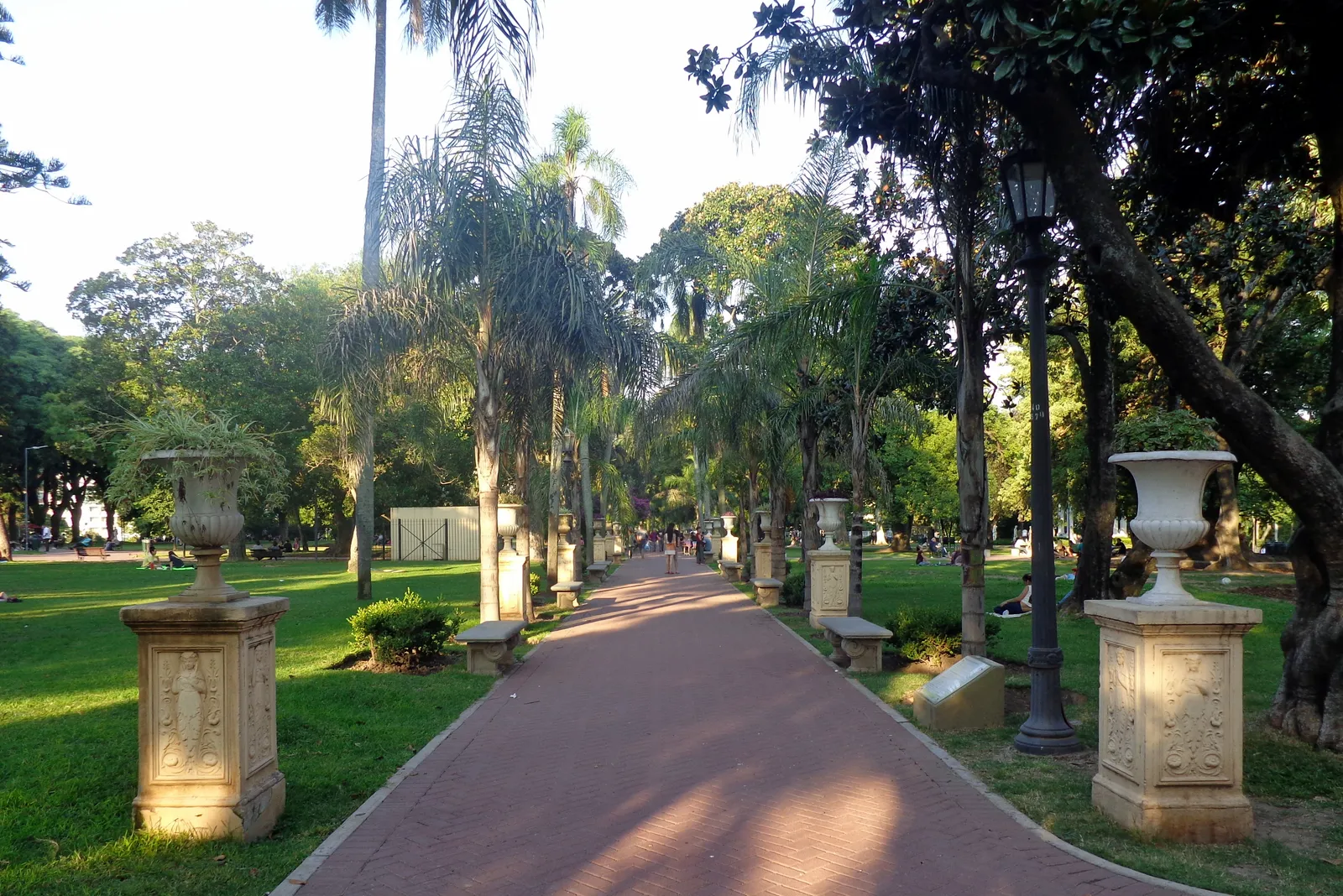A brick path through Parque Lezama in Buenos Aires, lined with tall palm trees and stone pedestals topped with decorative urns, with a wrought-iron lamppost beside the walk