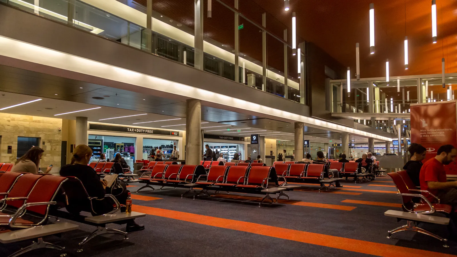 A gate waiting area inside Terminal B at Buenos Aires Ezeiza airport, with rows of red and grey seats on patterned carpet, shops along the back wall and a mezzanine above