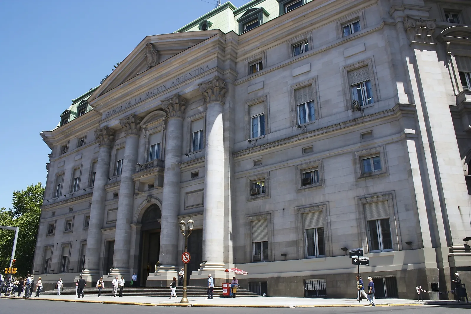 The columned Neoclassical entrance of the Banco de la Nación Argentina building on Plaza de Mayo in Buenos Aires, with tall stone pillars framing the doorway and pedestrians crossing the pavement in front