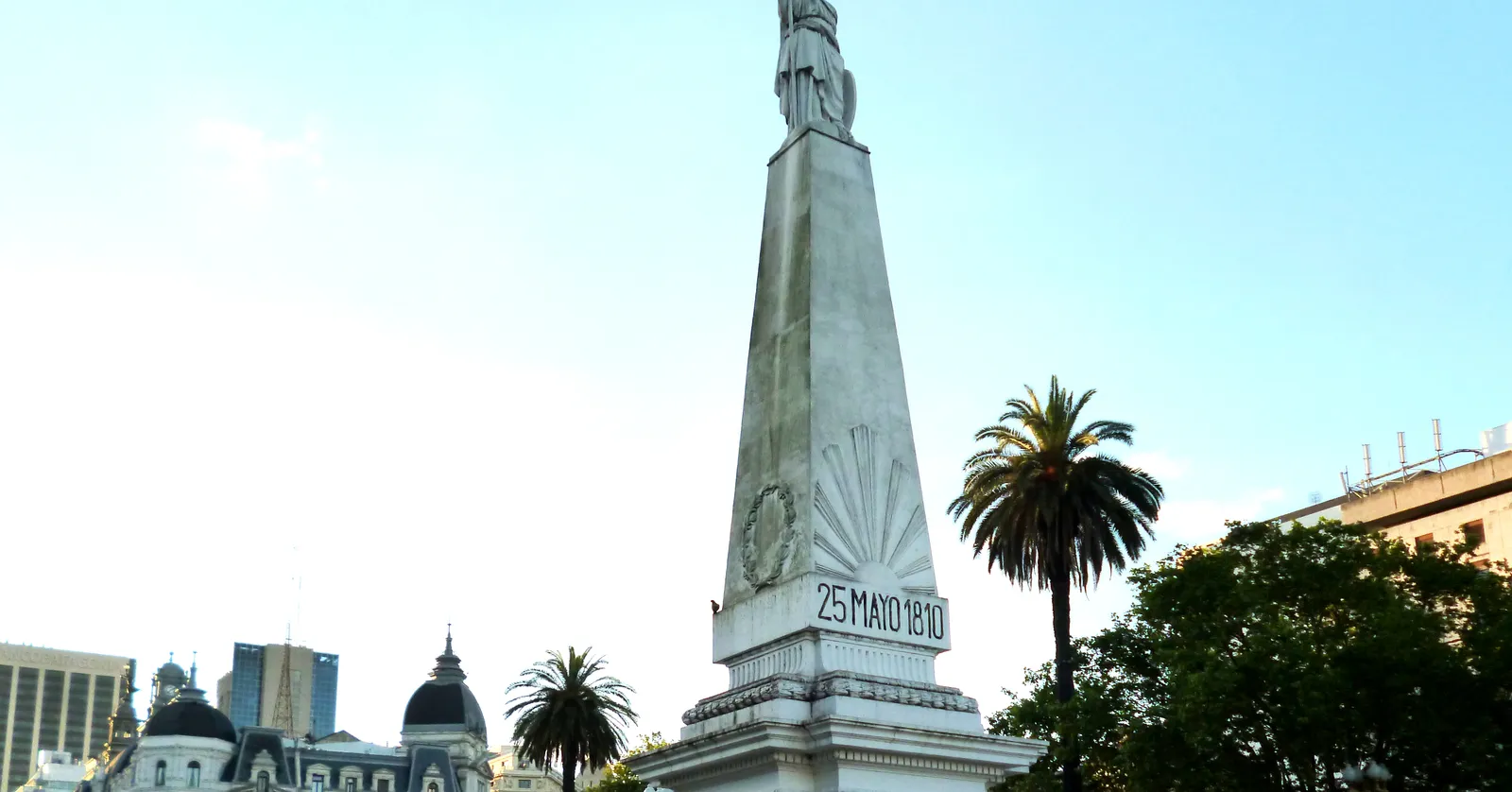 The white marble Pirámide de Mayo obelisk in Buenos Aires, topped with a statue of Libertad and inscribed 25 MAYO 1810, with a palm tree and the Cabildo's twin domes behind it