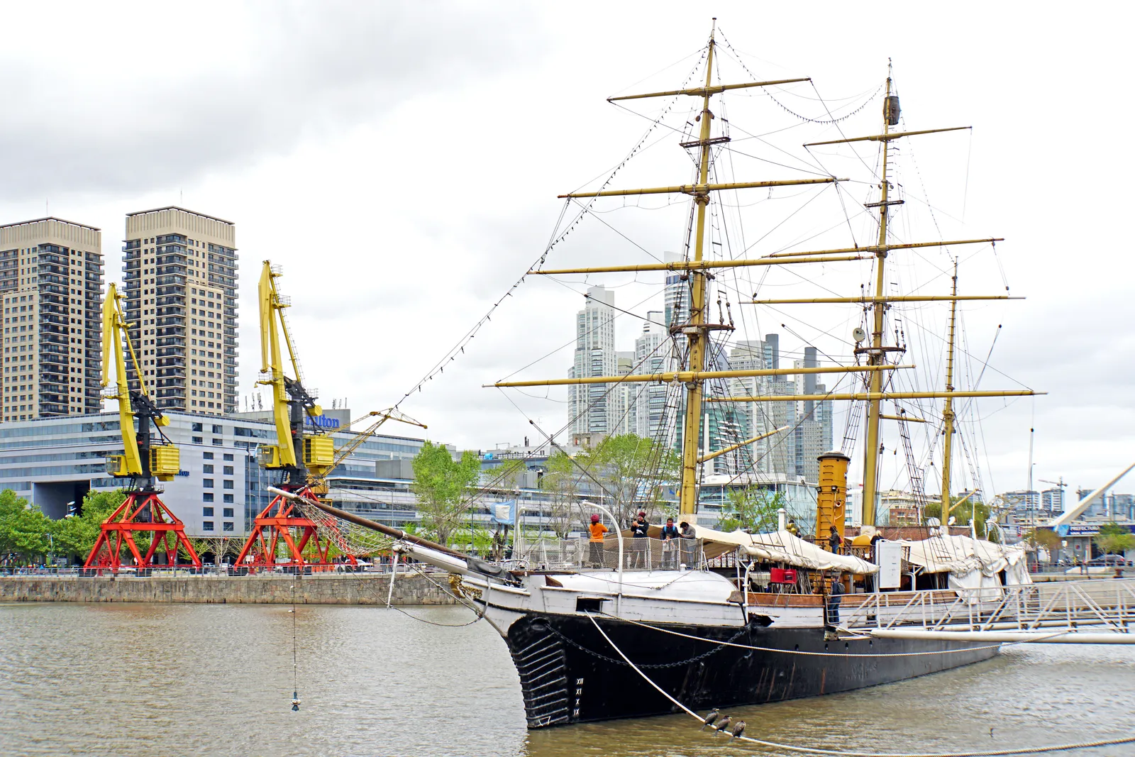 The black-hulled corvette Uruguay moored at a Puerto Madero dock in Buenos Aires, its three masts rigged and bowsprit projecting over the quay, with red harbour cranes and apartment towers behind
