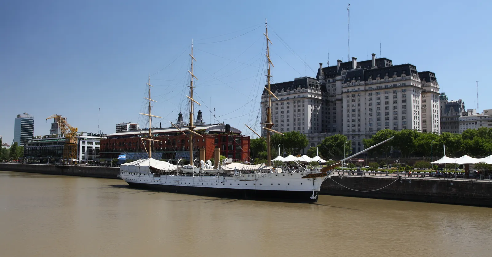 The white three-masted frigate Presidente Sarmiento moored on a dock in Puerto Madero, Buenos Aires, with red brick warehouses, a yellow port crane and a large stone government building on the quay behind it