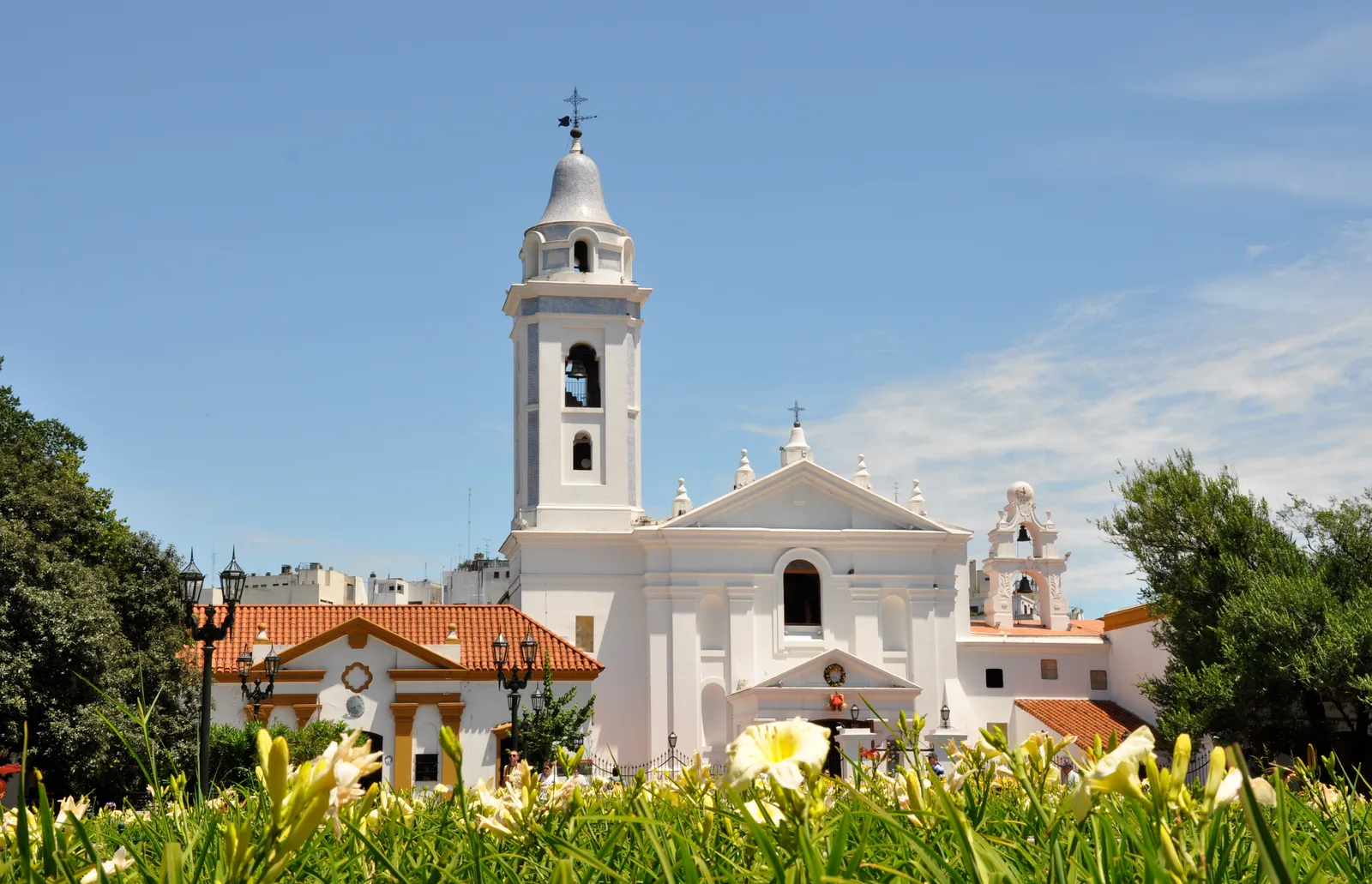 The whitewashed colonial front of the church of Nuestra Señora del Pilar in Buenos Aires, with a single domed bell tower, a tiled roof over the cloisters beside it and flowering shrubs in the foreground