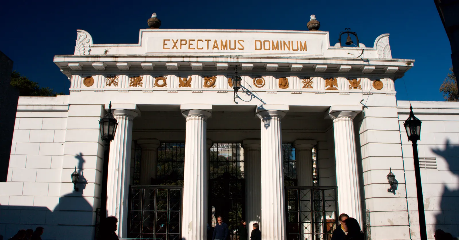 The white Doric portico at the entrance to Recoleta Cemetery in Buenos Aires, with gold medallions along the frieze, the words EXPECTAMUS DOMINUM above them and iron gates standing open between the columns