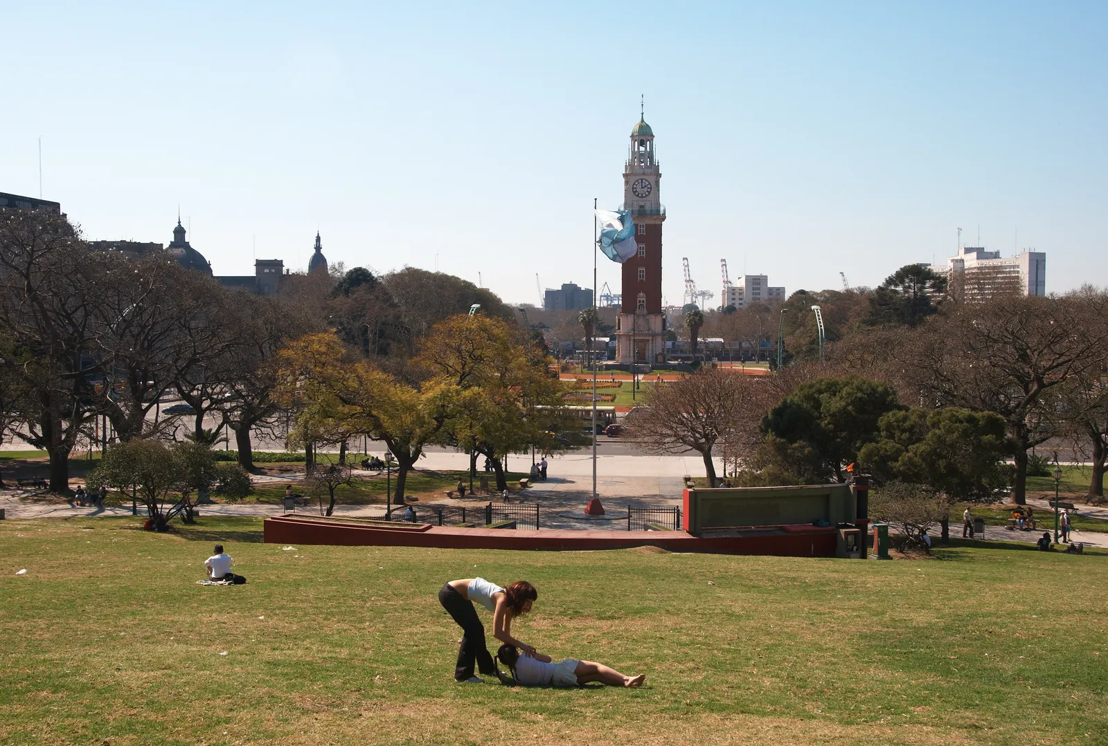 The sloping lawn of Plaza San Martín in Buenos Aires with people sitting on the grass under bare winter trees, and the clock tower and port cranes visible across the road at the bottom of the slope