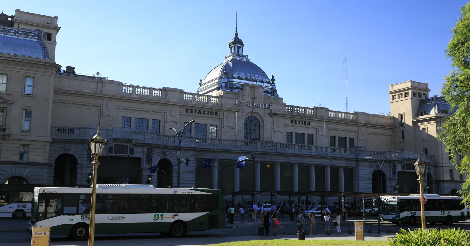 The long stone front of Retiro Mitre station in Buenos Aires with ESTACION RETIRO carved above the entrance, a green dome over the centre and city buses drawn up along the kerb