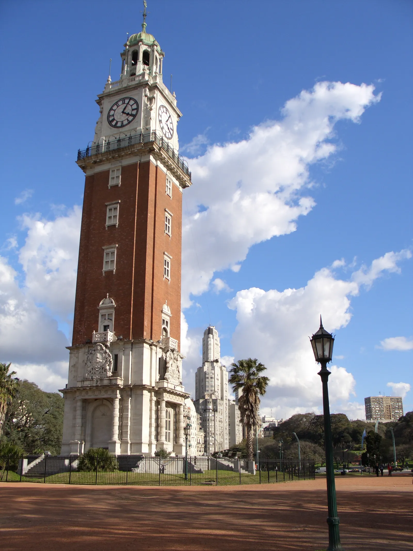 The red-brick Torre Monumental clock tower in Buenos Aires against a blue sky, its white stone base and belfry framed by palm trees, with the stepped white Kavanagh Building rising behind it