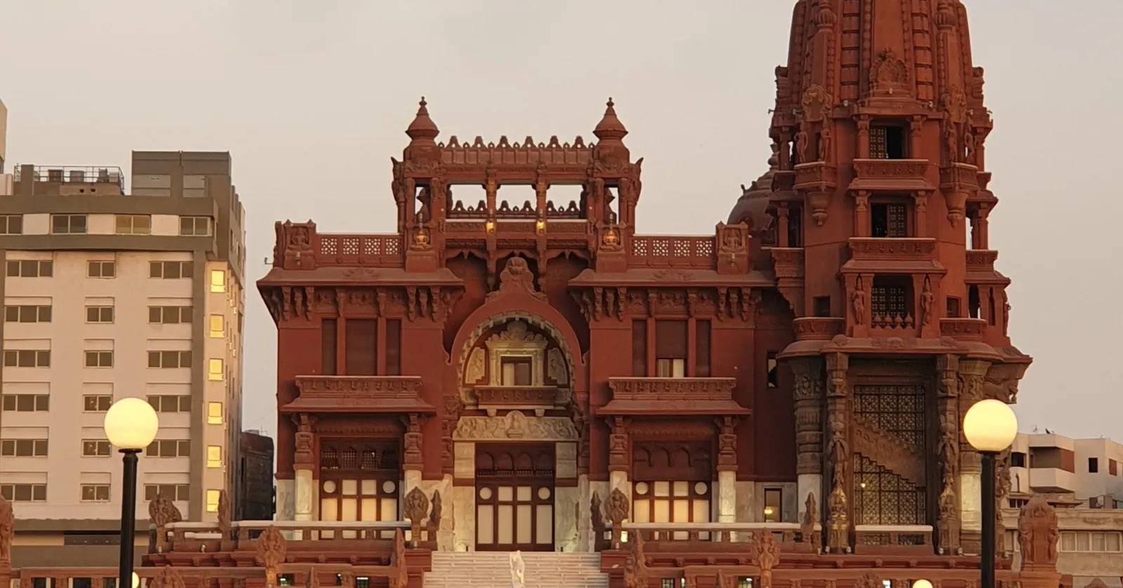 The restored Baron Empain Palace in Heliopolis lit at dusk, its deep red concrete facade carved with pillared galleries and temple turrets, modern apartment blocks standing behind it