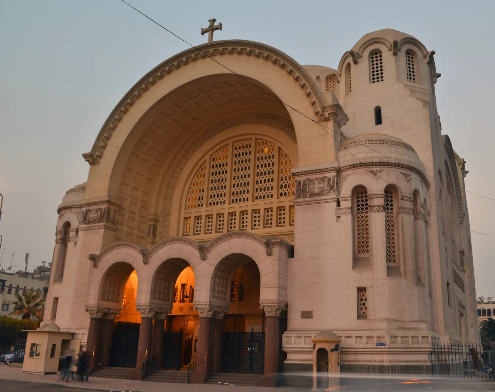 The Heliopolis basilica at golden hour, a huge recessed arch over the entrance arcade with a domed drum rising to the right and lamps lit under the porch