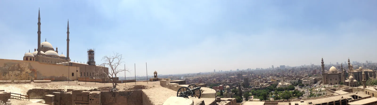 A wide view west from the ramparts of the Cairo Citadel, the domes and twin minarets of Muhammad Ali's mosque on the left, an old field gun on the wall, and the low rooftops of the city running to the haze