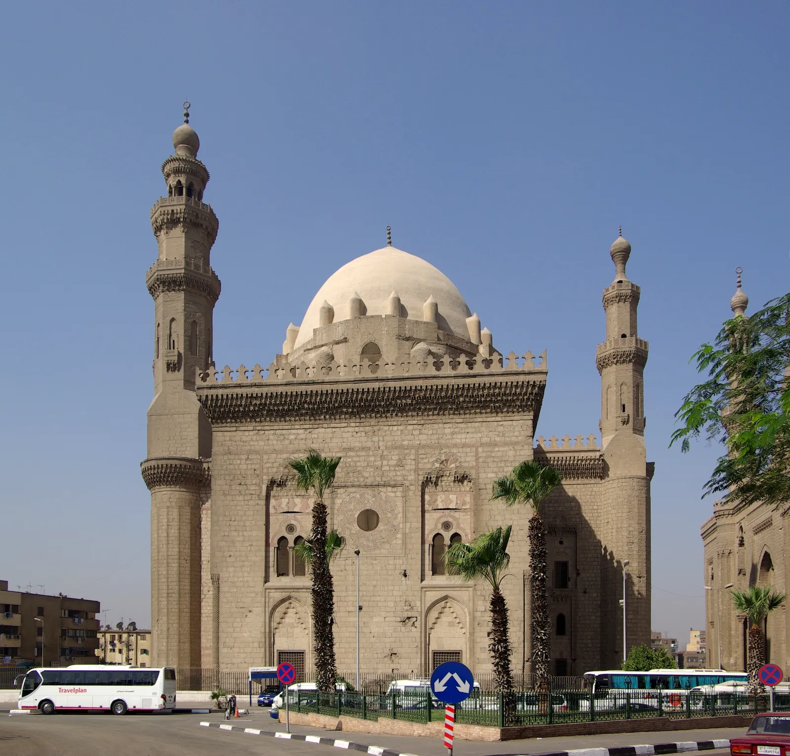 The stone bulk of the Sultan Hassan mosque and madrasa in Cairo, a tall blank-walled facade with a deep recessed entrance portal and a minaret rising from the corner