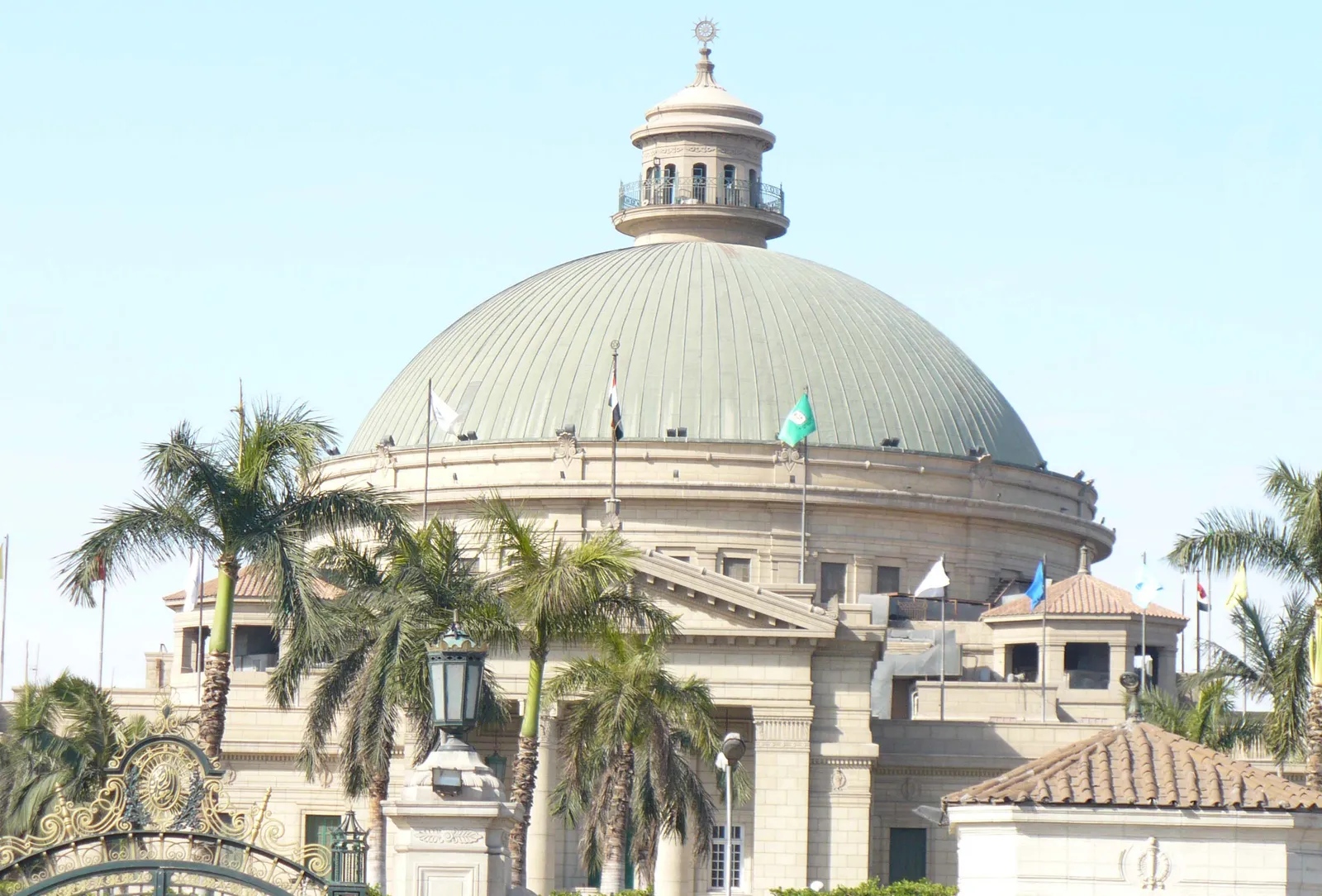 The green dome of Cairo University's main building rising above a stone colonnade, with palms and flagpoles in front of it and ornate iron gates at the bottom of the frame