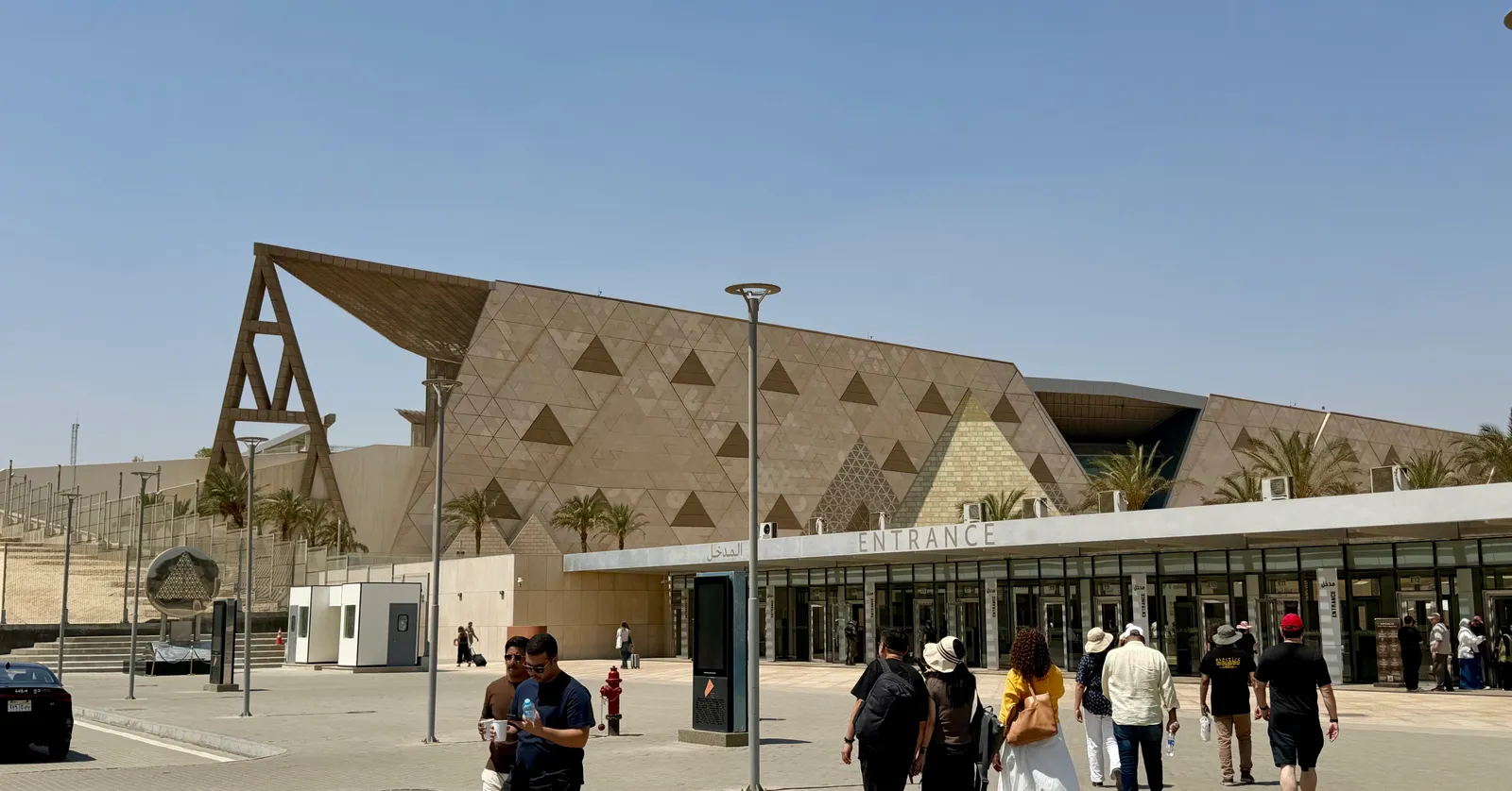 The long sandstone front of the Grand Egyptian Museum at Giza, its wall cut into a repeating pattern of triangles under a clear sky, with the glass entrance canopy and visitors crossing the forecourt
