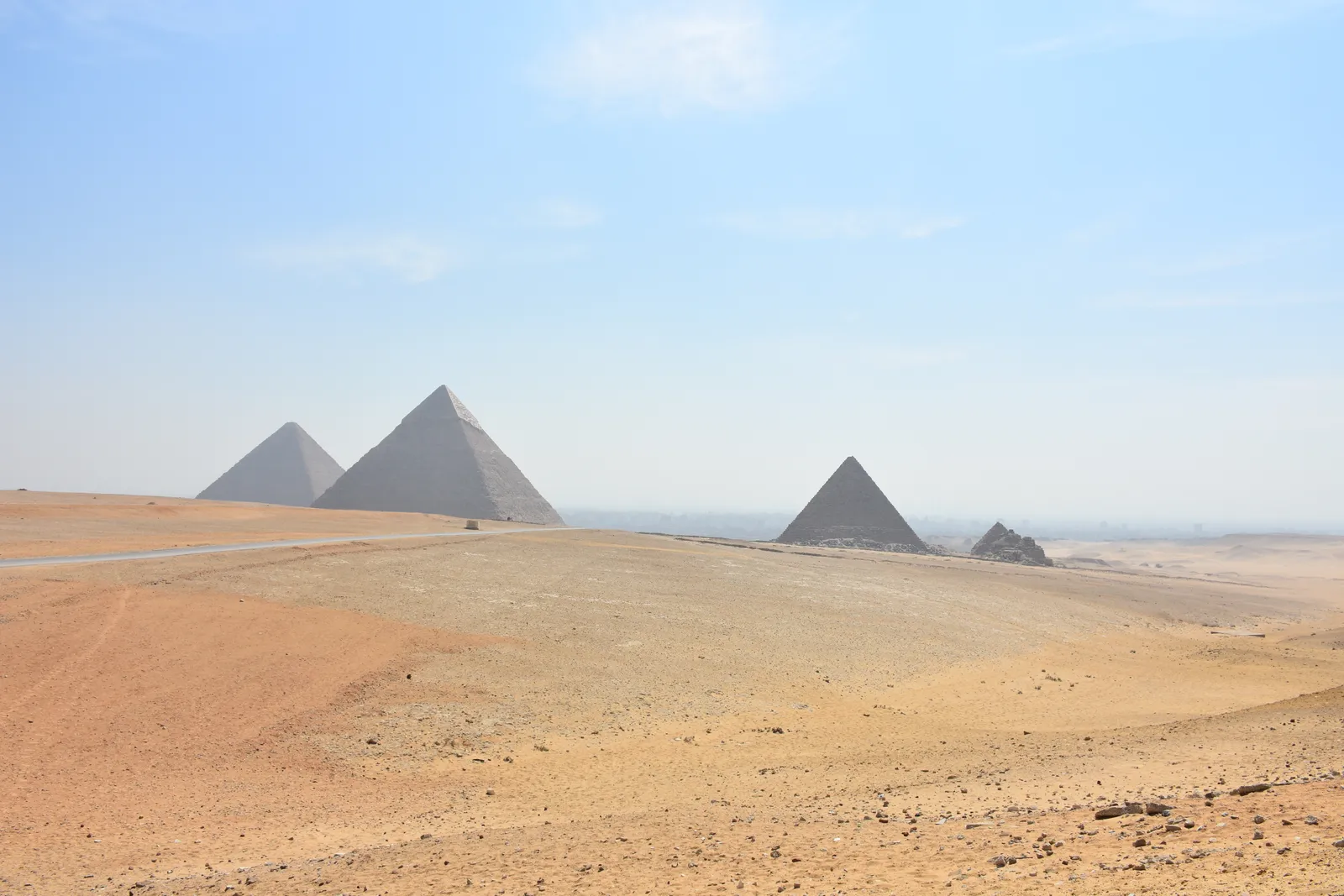The Giza pyramids seen from open desert to the south-west, the three large pyramids and the smaller satellite pyramids strung across the horizon above empty sand and a dirt track